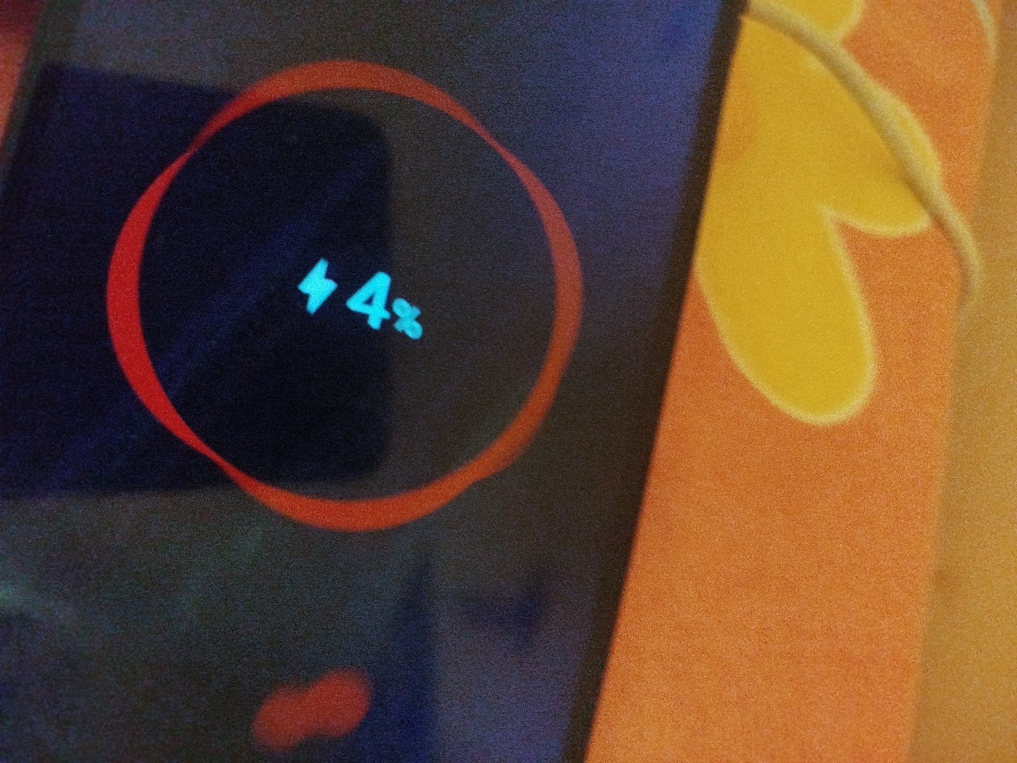 The image displays a close-up of a mobile phone screen showing a charging indicator. A red circle outlines a battery icon and the text "4%" in a bright cyan color. The phone screen is dark blue, and a blurry, out-of-focus object, possibly a charging cable, is visible in the upper right corner. The background is a textured surface with orange and yellow floral patterns. There is no information available in the image that would indicate its location in the city of Lassan, Pakistan.