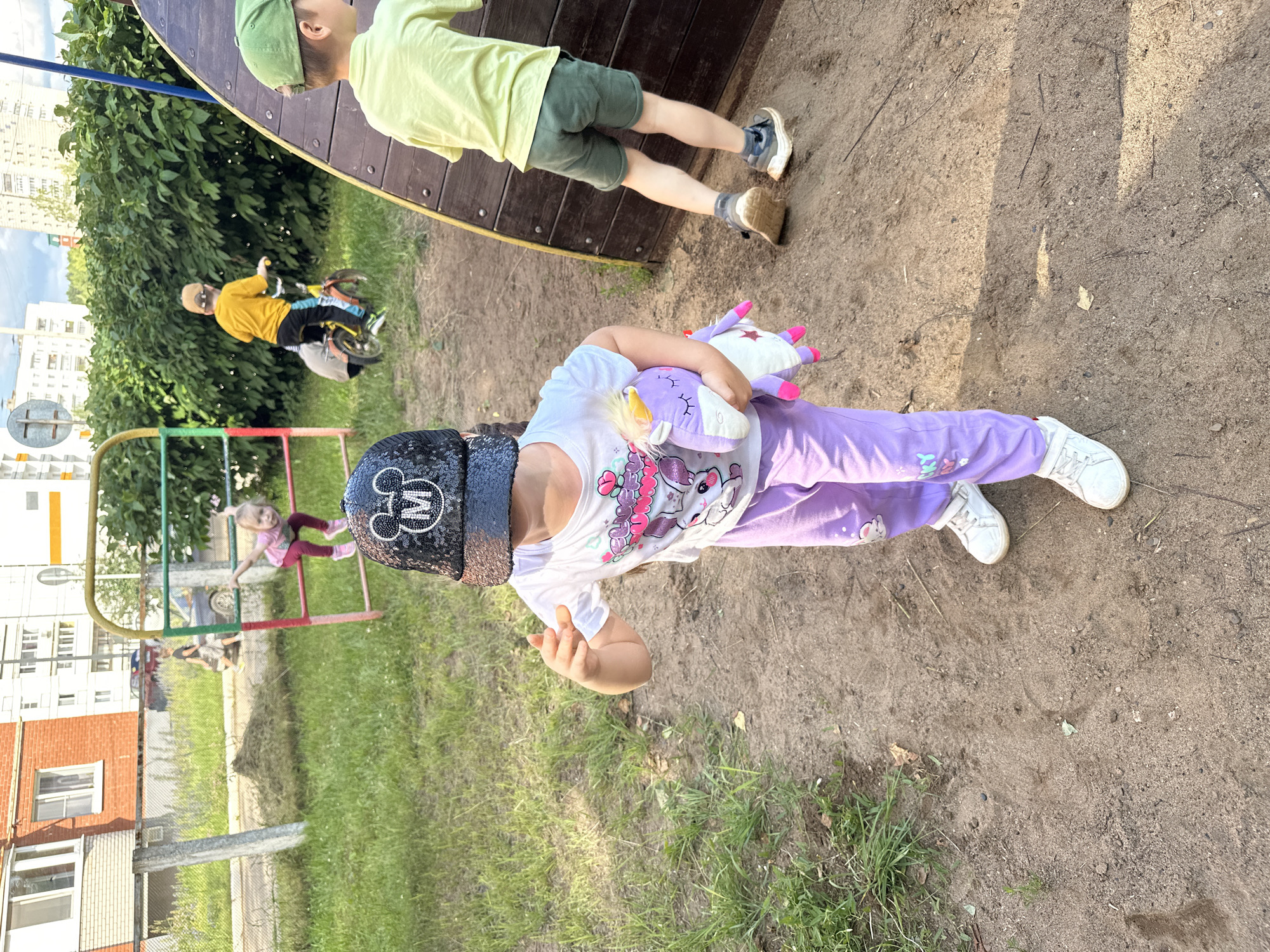 In a sunny outdoor scene, likely in Chaikovsky, Russia, a group of children are playing on a playground. A young girl in the foreground is wearing a white t-shirt with a unicorn design and purple pants, holding a stuffed unicorn. She is also wearing a black sequined hat with a Mickey Mouse design. To her left, a boy in a bright yellow shirt and blue shorts rides a small bicycle. In the background, another child in a light green shirt and dark green shorts is partially visible near a large wooden structure. A third child, a young girl, is on a red and green metal playground structure. The ground is a mix of sand and grass, suggesting a typical playground environment. The lighting indicates it is daytime, likely a warm day given the children's attire. The presence of apartment buildings in the background provides a clear indication of an urban or suburban setting. The overall mood appears to be one of carefree childhood play.