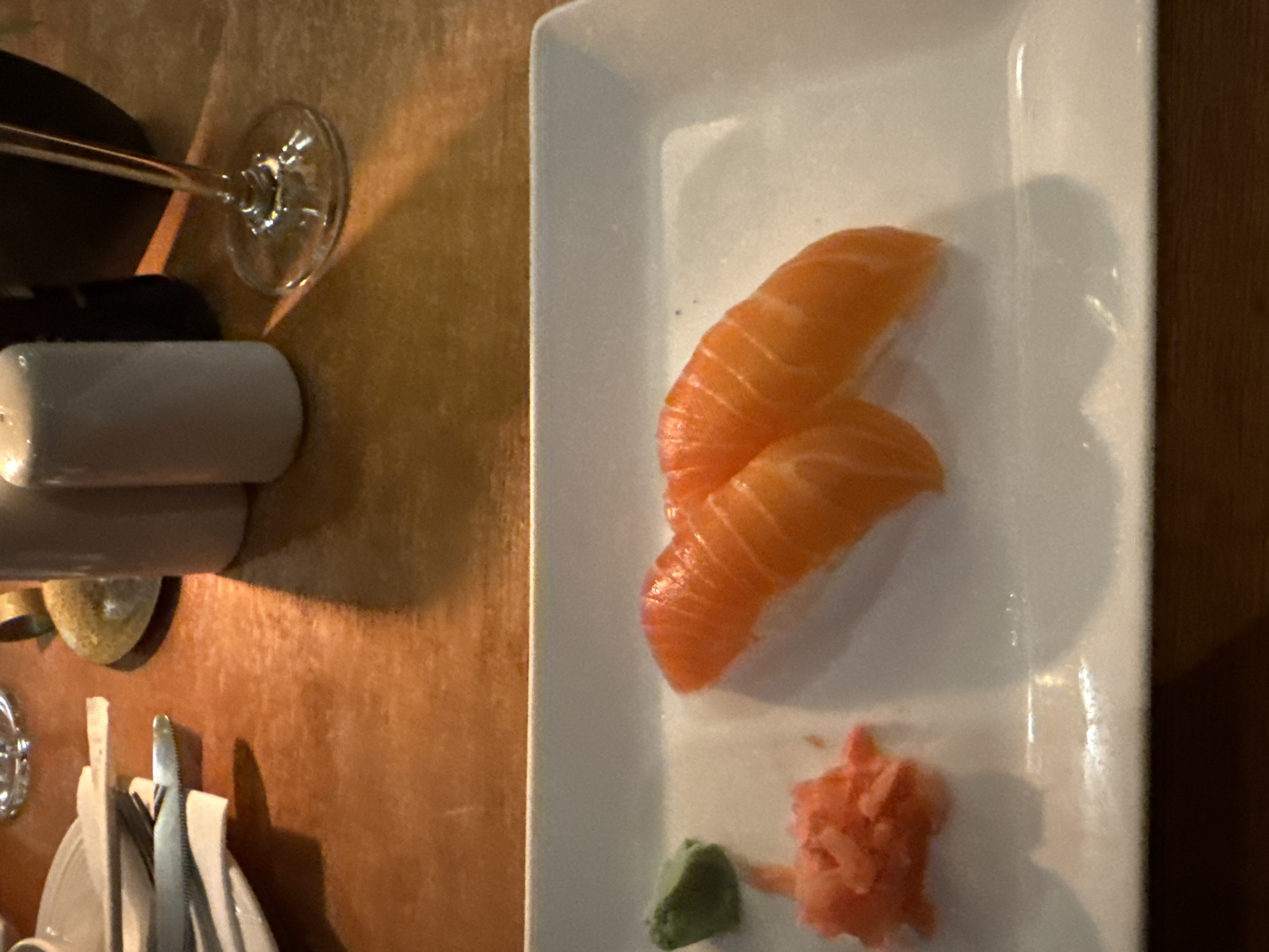Here is a detailed description of the image:
**Scene Overview:**
The image shows a meal presentation on what appears to be a wooden table, likely inside a restaurant. There is a rectangular white plate with salmon nigiri, wasabi, and pickled ginger. A wine glass is present on the table, partially obscured by a salt shaker. Cutlery can also be seen.
**Objects:**
* **White Plate:** Rectangular, holds the food.
* **Salmon Nigiri:** Two pieces of salmon over what is likely sushi rice.
* **Wasabi:** A small green mound.
* **Pickled Ginger:** Pink colored, shredded.
* **Wine Glass:** A stem glass, potentially containing wine.
* **Salt Shaker:** A cylindrical, white container.
* **Cutlery:** Appears to be forks and possibly a knife.
* **Table:** Made of wood
**Inferences and Context:**
* Given the presence of sushi, the location could be a Japanese or Asian-fusion restaurant.
* The presence of wine suggests a formal or upscale dining experience.
**Location:**
The image does not provide enough information to identify a specific location in Paarl, South Africa.