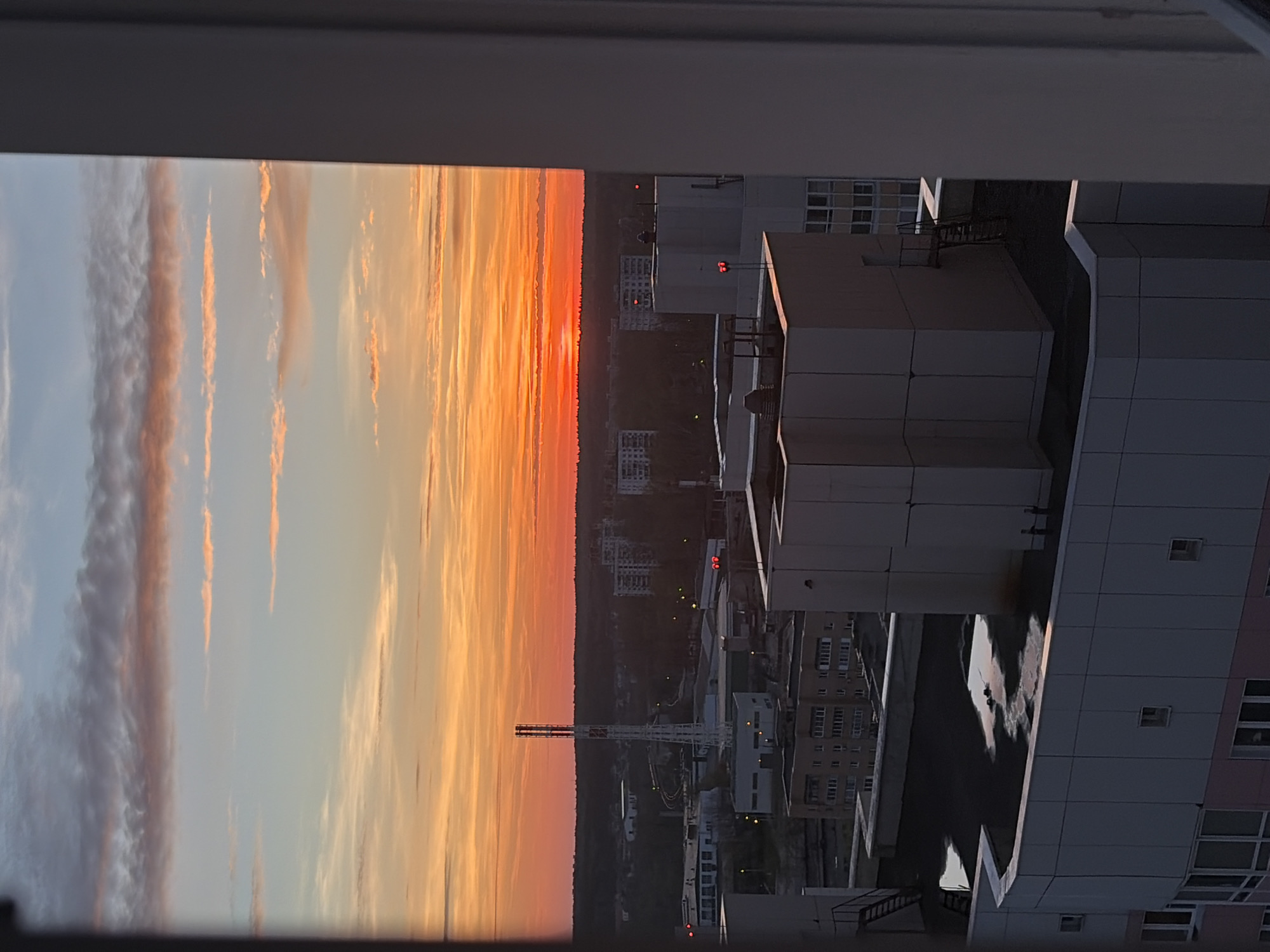 Here is a detailed description of the image:
**Setting:** An urban landscape observed from a high vantage point, likely an apartment window. The image captures a vibrant sunset sky with orange and red hues, contrasted by the structures of a city.
**Environment:** The sky features varied cloud formations illuminated by the setting sun. Below, the city includes a dense cluster of buildings, vegetation, and infrastructure. The overall atmosphere is one of dusk settling over a city.
**Objects:**
* Modern architectural buildings with rectangular structures and flat roofs.
* A tall industrial tower.
* Apartment buildings with numerous windows, some illuminated.
* Rooftop structures and possible access ladders.
**Actions/Interactions:**
* The sunset casts light on the city.
**Moscow, Russia Details:**
Although there are many cityscapes, some of the architectural elements (like the modern rectangular architecture) and the general layout of a dense urban area align with the characteristics that can be found in Moscow.