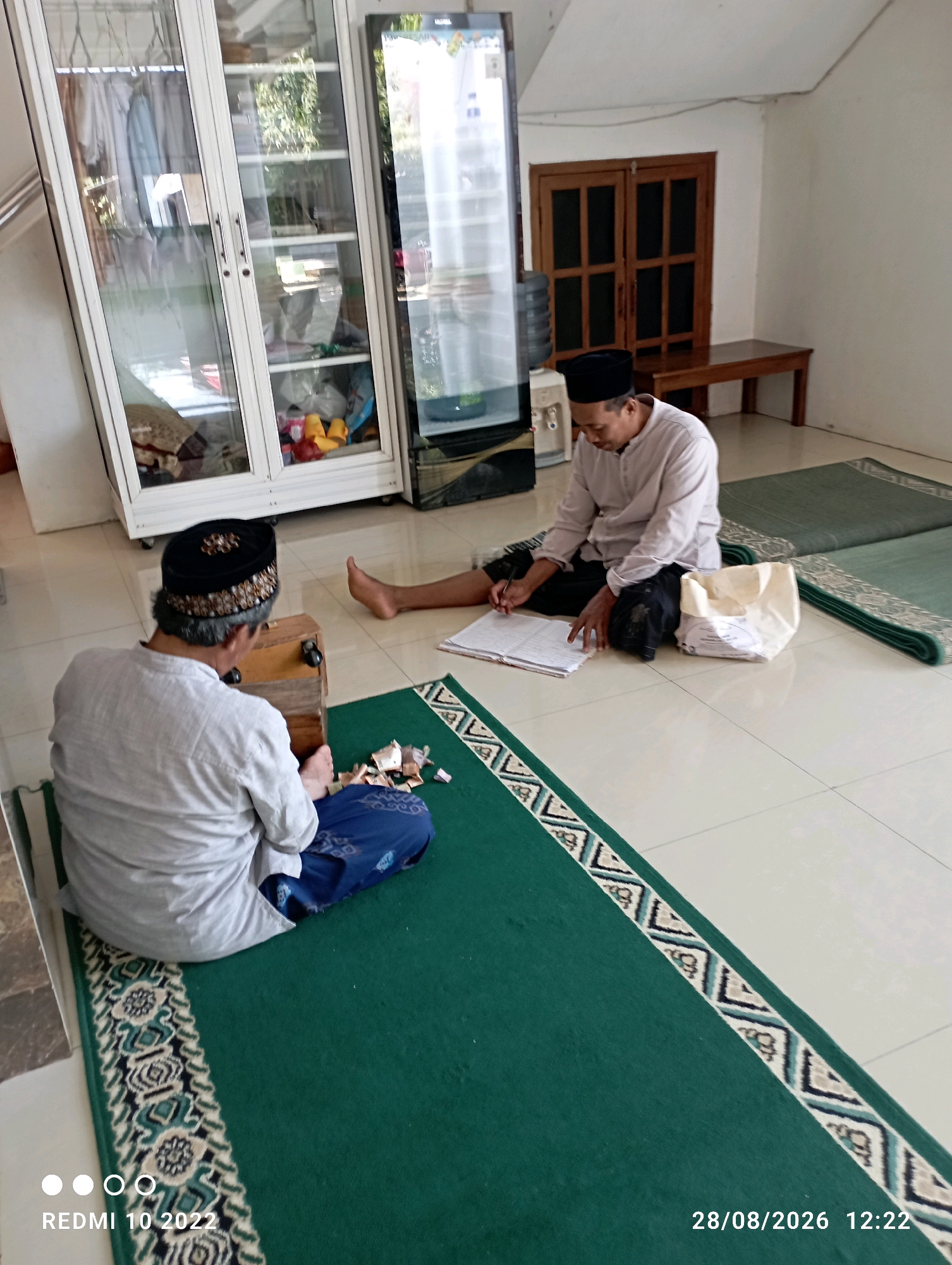 In a room in Dukuhseti, Indonesia, two men are seated on green prayer mats. The man on the left, wearing a white shirt and a black and gold patterned cap, is facing away from the camera. He is holding a wooden box with a metal latch and has papers scattered around him. The man on the right, wearing a light pink shirt and a black cap, is facing left, looking down at a notebook and pen, with a white cloth bag beside him. The room has tiled flooring and white walls. A tall, white cabinet with glass doors, a black refrigerator, and wooden doors with a bench are visible in the background. The scene appears to be indoors, possibly in a mosque or a similar community space.