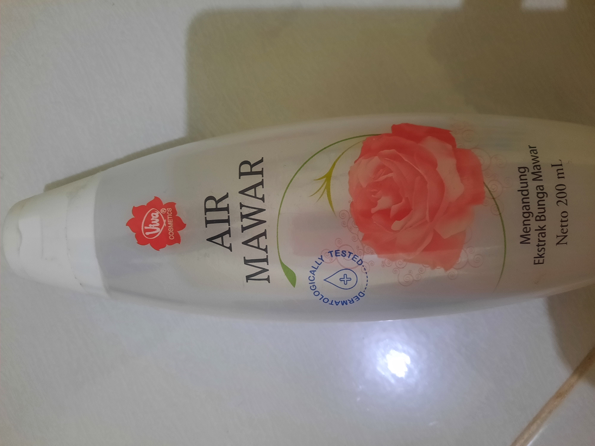 A clear plastic bottle of Viva Air Mawar skincare product is displayed upright against a tiled, possibly bathroom, surface. The bottle is oval-shaped with a white cap and contains a clear liquid. The label features a red Viva Cosmetics logo, the text "AIR MAWAR" in large black letters, a floral illustration of a pink rose, and a circular seal indicating "DERMATOLOGICALLY TESTED" with a droplet icon. Below the rose illustration, the text reads "Mengandung Ekstrak Bunga Mawar" and "Netto 200 mL". The background is a light-colored, textured tile. There are no people or animals present in the image. The lighting suggests an indoor environment, potentially a home or commercial establishment in Lhokseumawe, Indonesia.