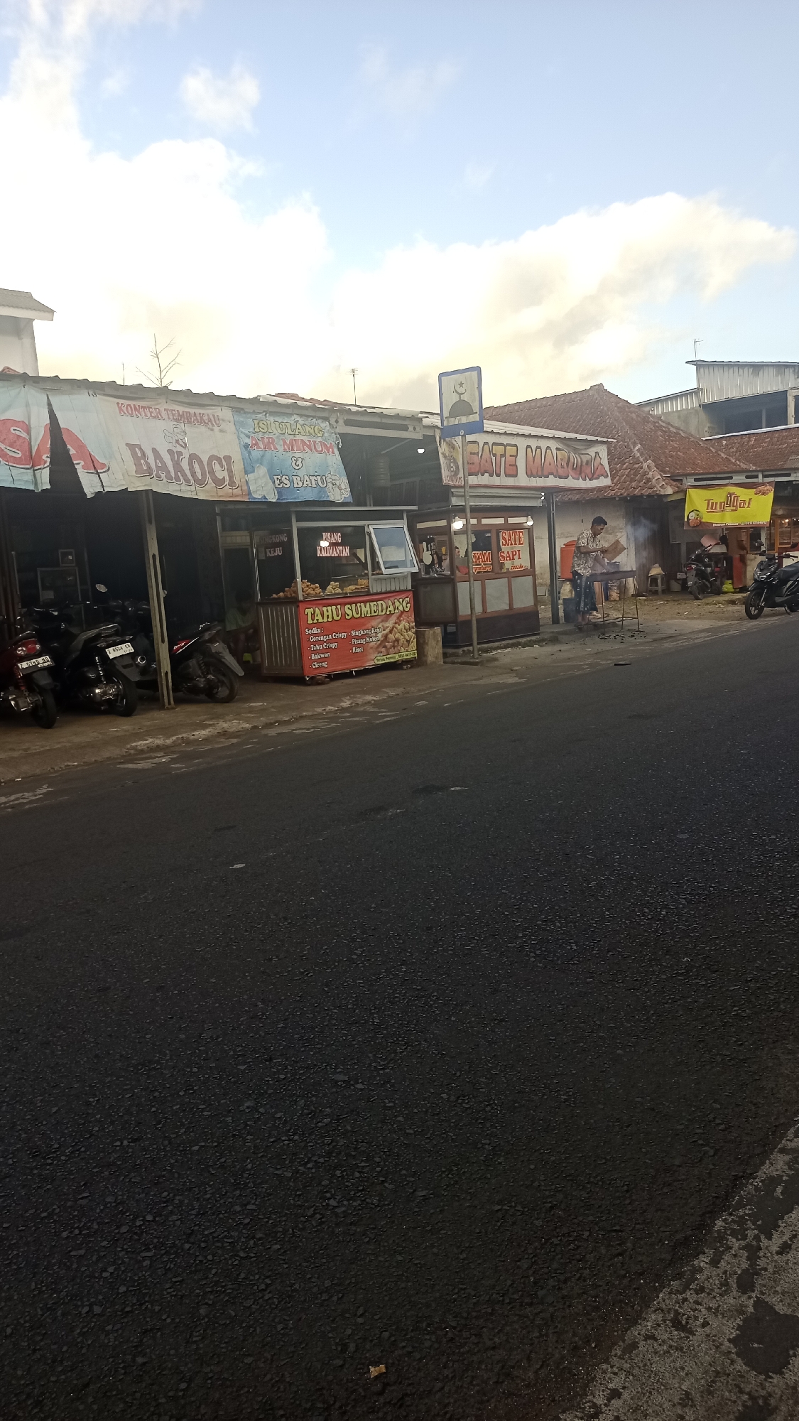 The image shows a street scene in Pakemitan, Indonesia, with several food stalls lining the roadside. On the left, there are motorcycles parked in front of a stall with banners advertising "BAKOCI" and "KONTOR TEMBAKU". Further down, there is a stall selling "TAHU SUMEDANG", with a menu visible listing various fried items. To the right of the Tahu Sumedang stall, a man is seen tending to a grill at a "SATE MADURA" stall. In the background, more food stalls and buildings are visible, with a bright sky and clouds overhead. The road in the foreground is asphalt and appears to be wet in places. The overall atmosphere suggests a bustling local market or street food area.