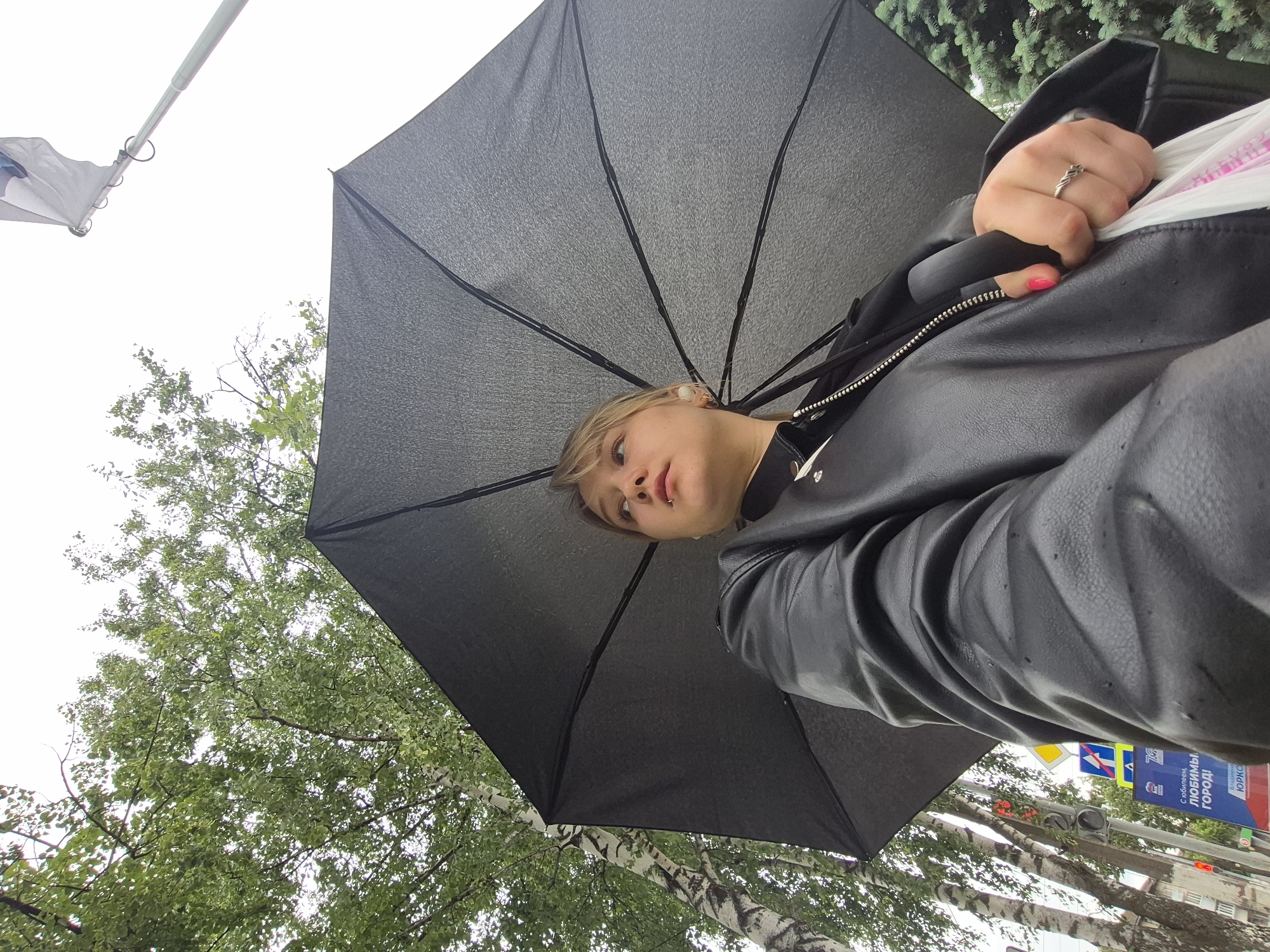 A low-angle photograph captures a young woman holding an open black umbrella outdoors in Chaikovsky, Russia, during overcast conditions. The camera is positioned beneath the umbrella, looking up at her and the sky. The woman has light-colored hair, fair skin, and a small stud or piercing above her upper lip. She is wearing a black leather-like jacket with a zippered front and has white wireless earbuds in her ears. On her right hand, which has pink nail polish, she wears a silver-colored ring and is holding a white, possibly plastic, item.
The sky is a bright, uniform grey, indicating an overcast day, and the presence of the open umbrella suggests it may be raining or has recently rained. Lush green foliage from various trees frames the scene, including deciduous trees with distinctive white birch bark and some coniferous trees.
In the background, street elements are visible, including a prominent blue sign with Russian text. The sign reads: "С юбилеем, любимый, город!" which translates to "Happy Anniversary, beloved city!" Below this, partially visible, is the word "ЮРКО". A yellow marker displaying the number "75" is also present, likely related to the anniversary mentioned on the sign. A flagpole with a white flag or banner is visible against the sky in the upper left corner. The woman's gaze is directed upwards towards the camera, and her expression is neutral, capturing a moment from what appears to be an urban outdoor setting during a local celebration.