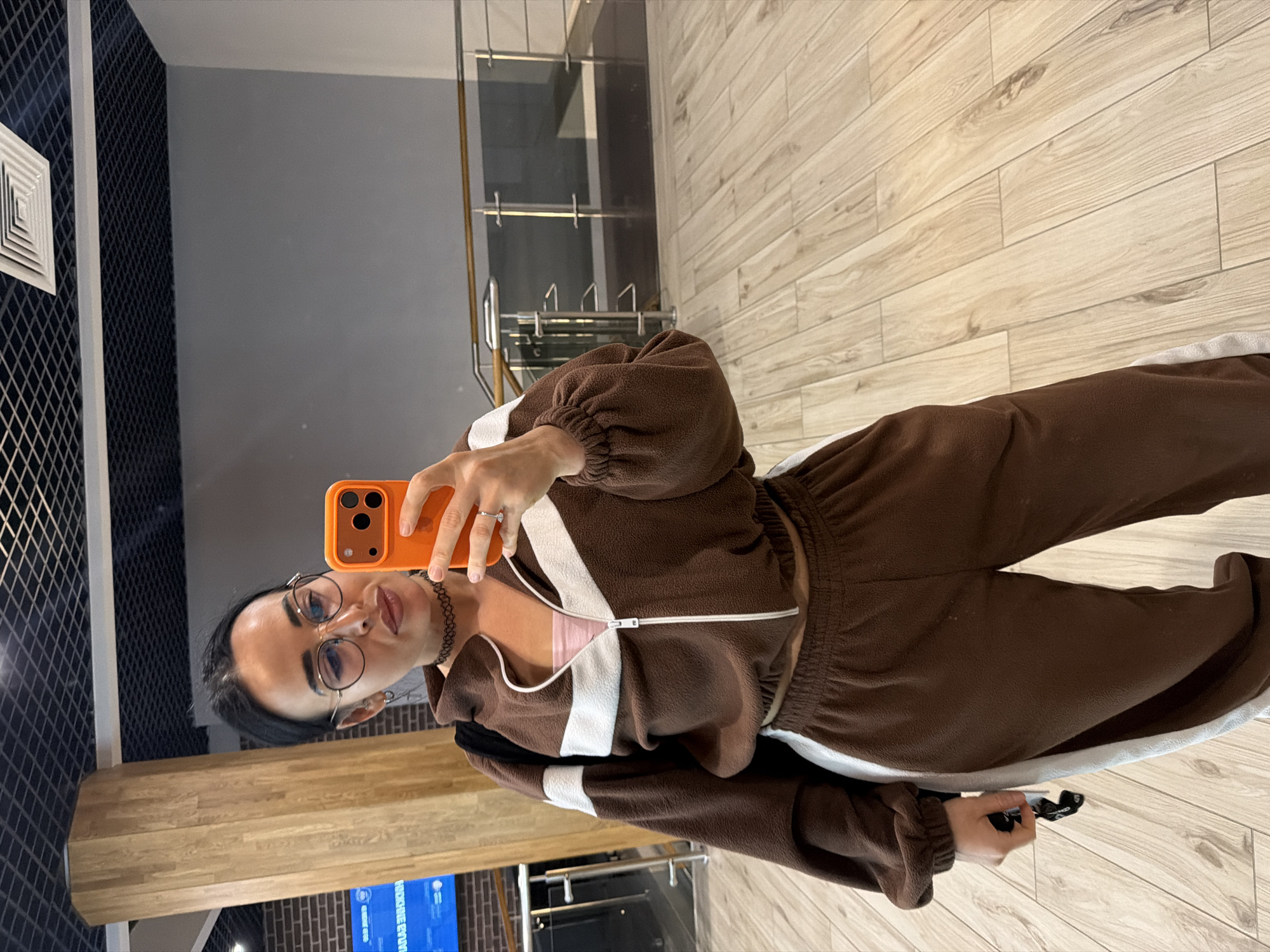 The image displays a young woman taking a mirror selfie indoors. She has dark hair, wears round eyeglasses, and a black choker. Her outfit consists of a casual, long-sleeved brown fleece tracksuit with white stripes on the chest, sleeves, and pants, and a light pink top visible underneath her zipped-up jacket. She holds an orange-cased smartphone in her right hand to capture the reflection and a small black object, possibly a bag or clutch, in her left hand. A ring is visible on her right ring finger.
The setting appears to be a modern indoor environment, characterized by light wooden plank flooring, a mirrored wall in which the selfie is taken, and a dark patterned wall with a white square air vent to the left. In the background, a glass railing and what seems to be a staircase or elevated platform are visible. The bottom portion of the reflection shows a brick wall and a display screen with indistinct blue and white text. The lighting suggests an indoor, well-lit space. The woman maintains a posed, neutral expression. The entire image is oriented 90 degrees clockwise from typical portrait view.