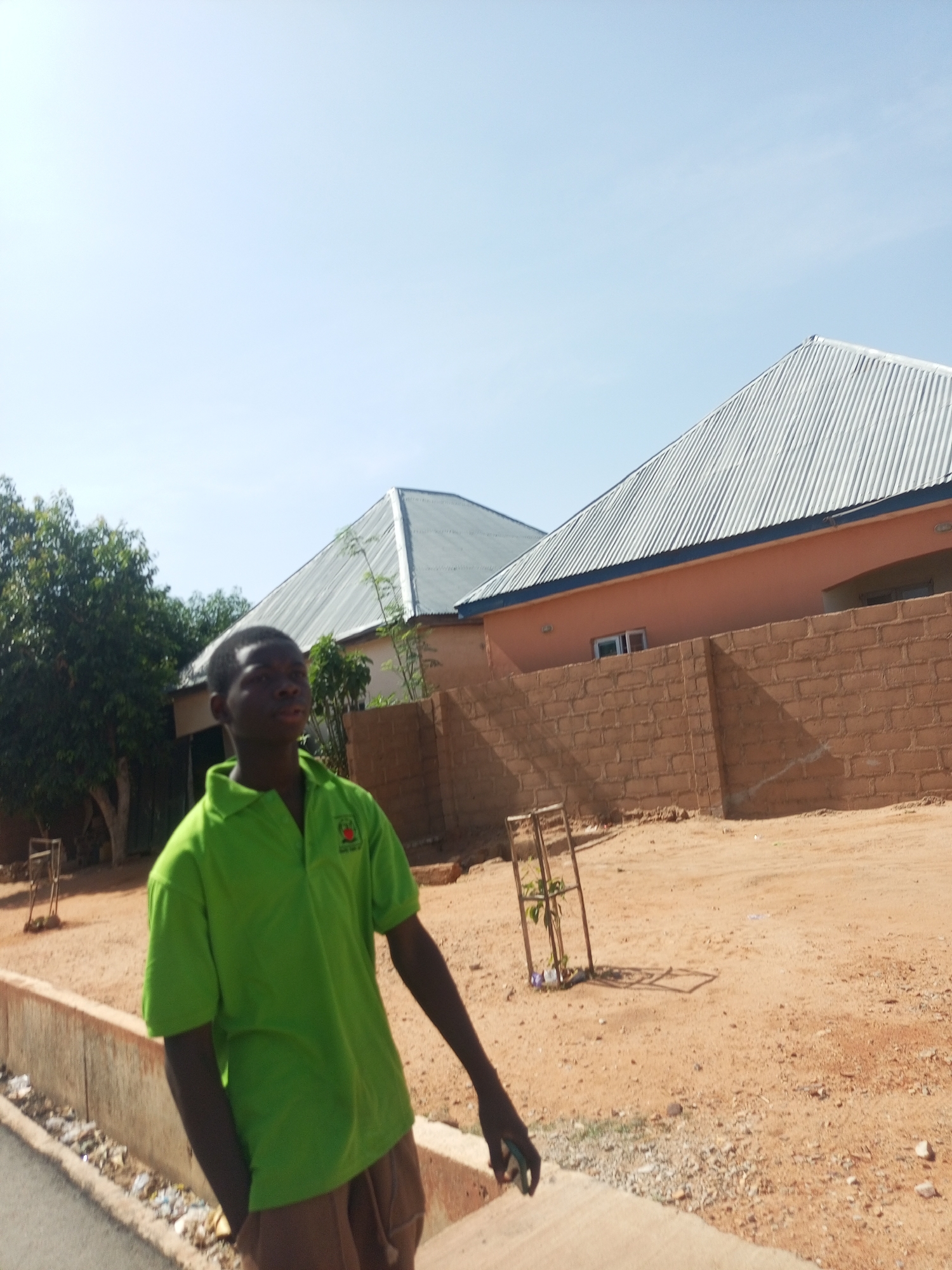 The image depicts a young man standing on a sidewalk in a residential area of Unknown, Unknown. He is wearing a green polo shirt and brown pants, with one hand in his pocket. Behind him are two single-story houses with grey, corrugated metal roofs and light brown walls. A brick wall encloses the houses. The ground is sandy, and a small, fenced-in tree is visible in the foreground. A leafy tree is to the left of the man. The sky is clear and light blue.