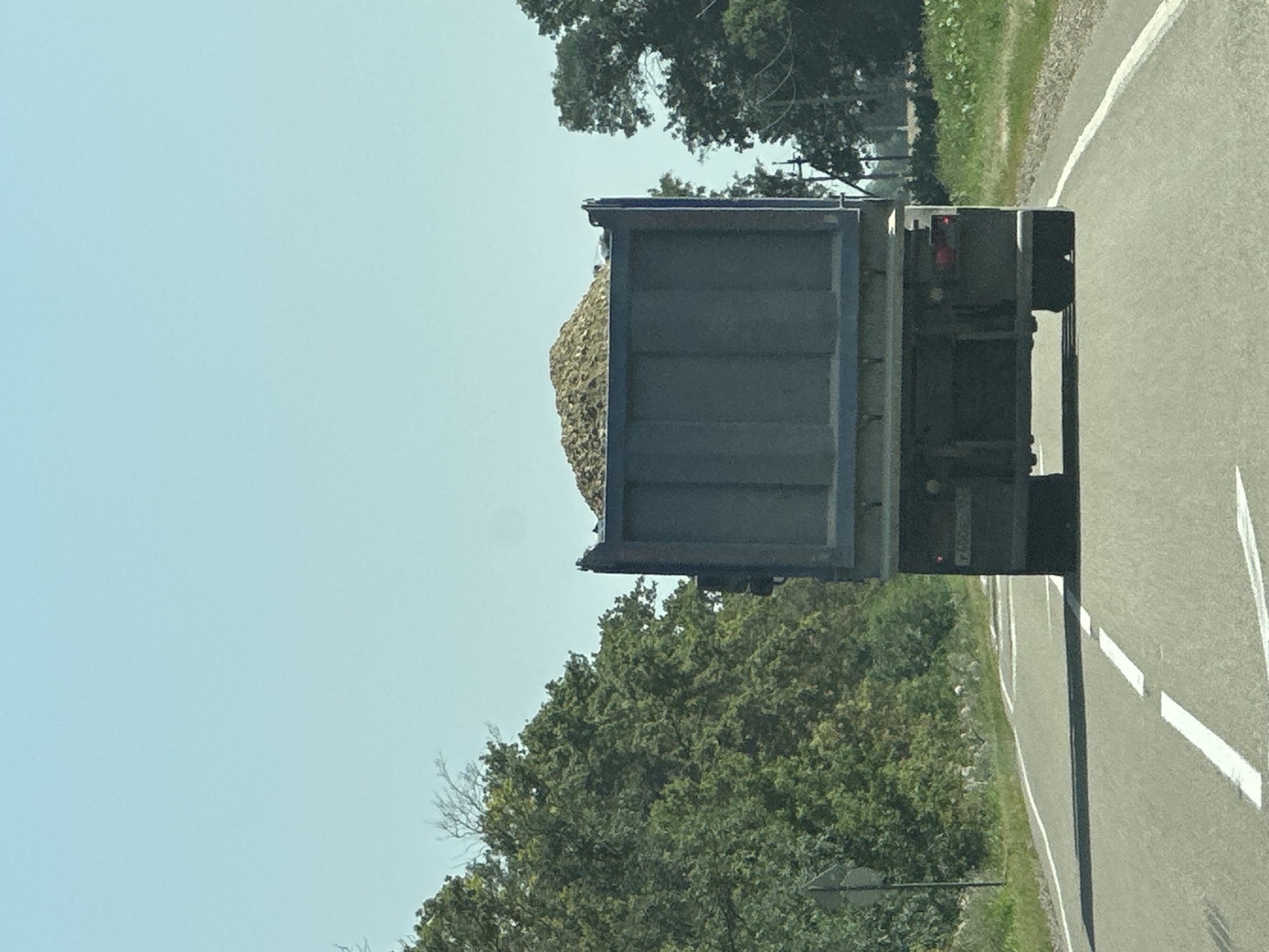 A rear view of a large, dark blue dump truck filled with light-colored gravel is traveling on a paved road. The truck's brake lights are visible, and it has dual rear wheels. The road has white dashed lane markings and a solid white line on the right edge. To the left of the road are dense green trees and a road signpost. To the right of the road is a grassy embankment with some vegetation. The sky is clear and blue. The image appears to be taken from another vehicle on the same road. The location is Tysyachny, Russia.