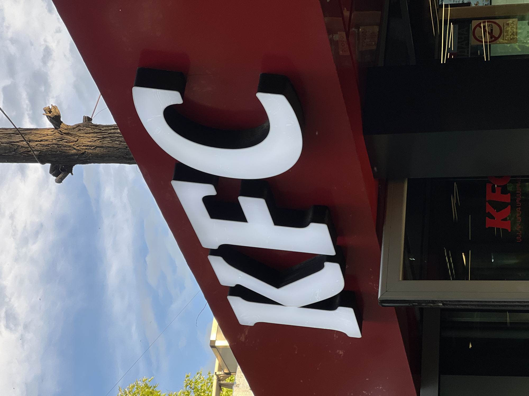 The image shows a KFC restaurant sign in Yerevan, Armenia. The letters "KFC" are displayed vertically on a red background. The letters are white with black outlines. A utility pole is visible in the background, and there are glass windows with visible signage to the right of the KFC sign. The sky is visible in the upper-left corner.