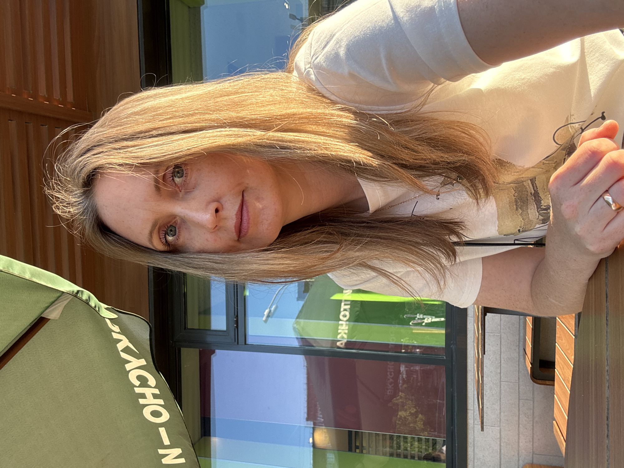 A female individual is positioned in the foreground, seated at an outdoor wooden slatted table, facing the camera. She possesses long, straight, light-brown hair, blue eyes, and freckles visible on her face and left forearm, which rests on the table. She wears a cream-colored short-sleeved t-shirt with a faint graphic design on the chest and a simple ring on her left ring finger.
Behind her, a modern building with a glass facade is visible. The building's exterior features vertical panels in shades of green and maroon/red. Reflected in the glass windows, the Cyrillic word "ВКУСНО" (VKUSNO), meaning "delicious," is clearly legible, identifying the establishment. A large green outdoor umbrella, partially visible in the upper left, also displays inverted and reversed Cyrillic text, which, in conjunction with the reflection, confirms the location as "Вкусно - и точка" (Vkuno - i tochka), the Russian successor to McDonald's. Additional reflections in the windows show parts of the surrounding urban environment, including a street lamp and other structures. The scene is illuminated by bright, natural sunlight, casting warm tones and creating distinct highlights on the subject and immediate surroundings. The location is specifically identified as "Вкусно - и точка" at Pushkinskaya Street, 268, in Izhevsk, Russia.