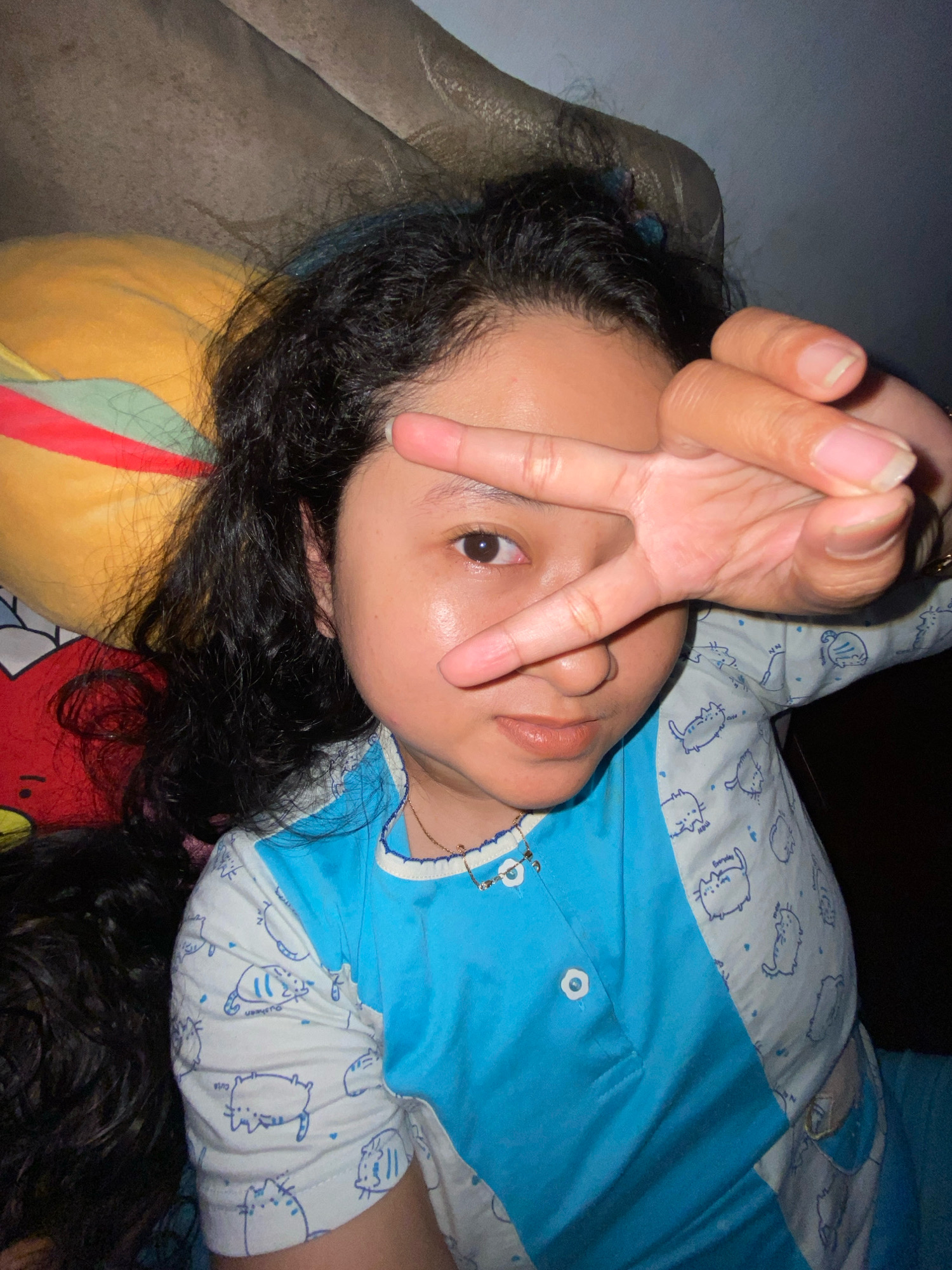 A young female with dark, curly hair lies on her side, propping her head on a pillow. She is wearing a blue and white long-sleeved shirt with a pattern of cartoon cats. Her right hand is raised, with her index and middle fingers extended in a peace sign, covering part of her face. The background appears to be a dimly lit indoor setting, possibly a bedroom, with a couch and a pillow visible. The image is a close-up, focusing on the subject's face and upper body. No specific location in Gelam, Indonesia is discernible from the visual information.