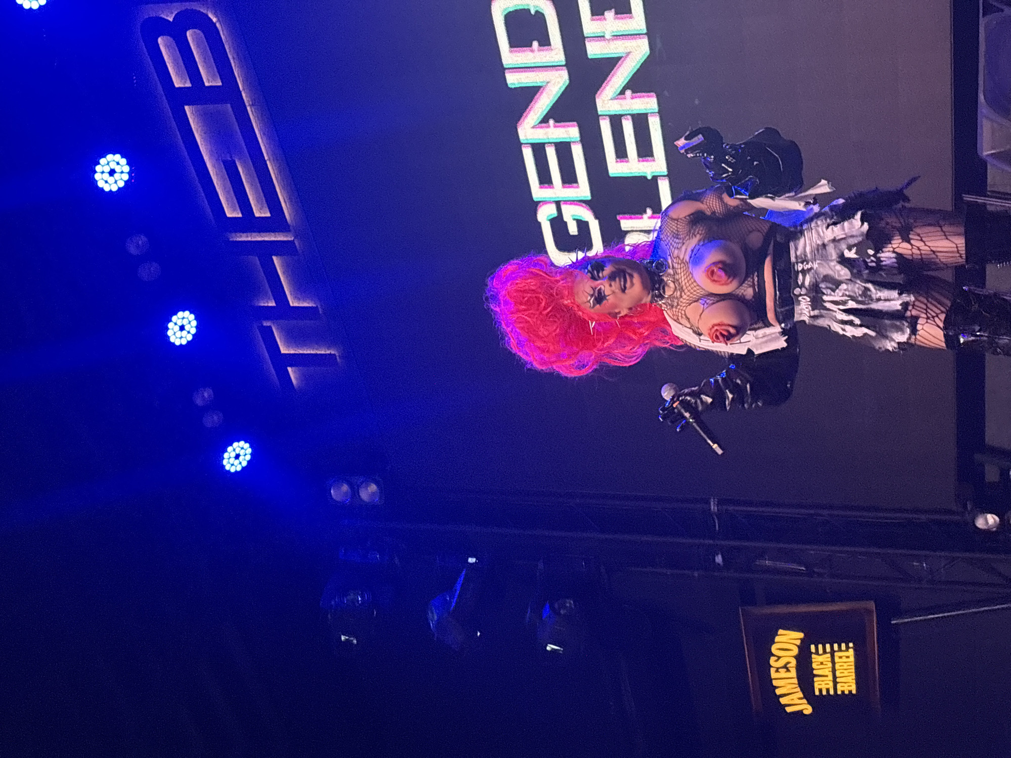 A drag performer with bright pink hair and dramatic makeup stands center stage, holding a microphone and wearing a revealing outfit consisting of a fishnet top, a black and white distressed skirt, and thigh-high boots. The performer has prosthetic breasts with nipple coverings. Behind the performer, a large screen displays the words "THE B" and "GENDER BLENDER" in large, illuminated lettering. To the left, a lit sign for "JAMESON BLACK BARREL" is visible. The lighting is predominantly blue, with spotlights and stage lights illuminating the scene. The setting appears to be an indoor venue in Varna, Bulgaria, likely a club or theater hosting a performance.