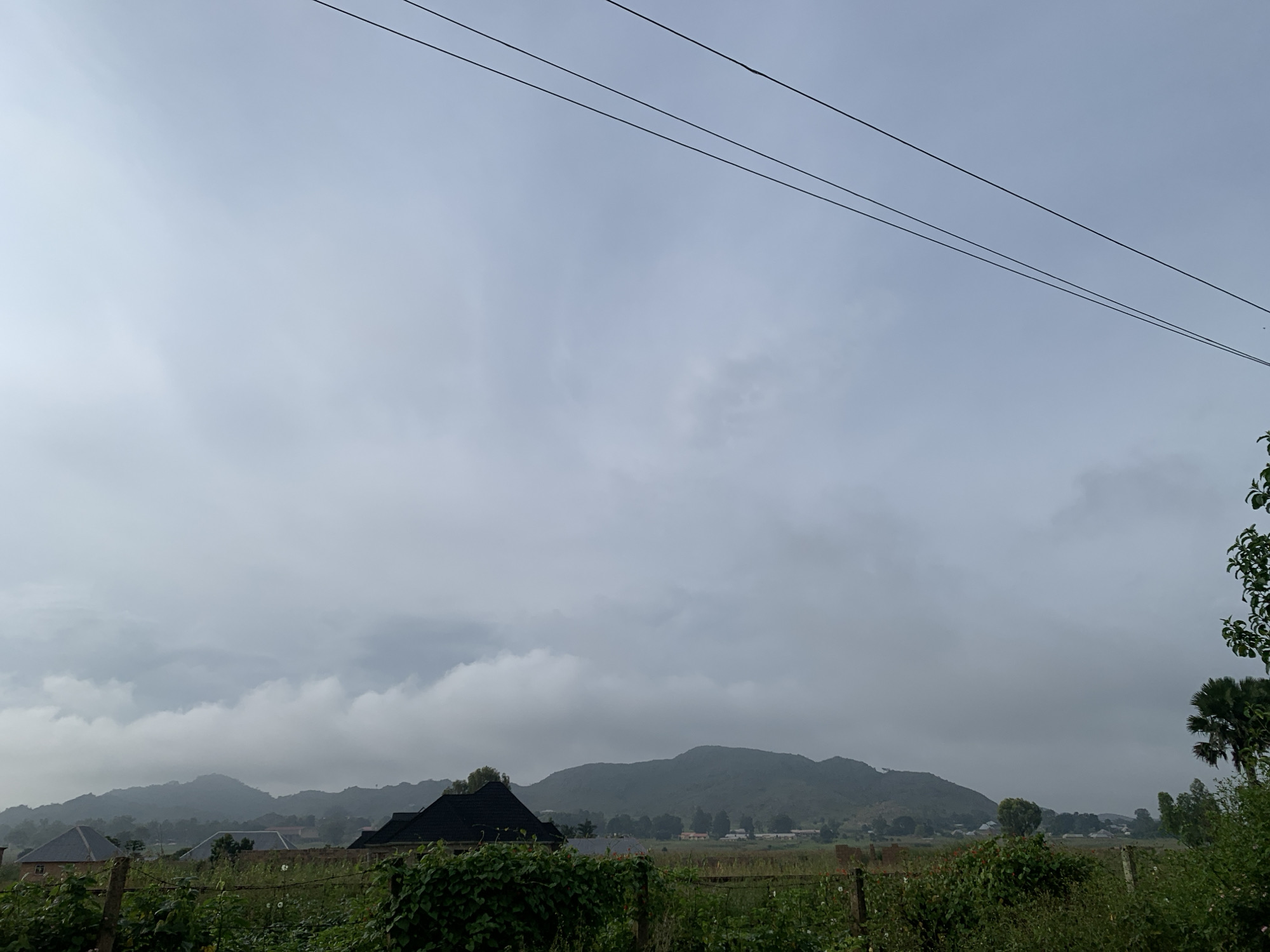 A landscape scene shows a cloudy sky over a rural area. In the foreground, there is dense green foliage and a wooden fence. Beyond the foliage, several houses with dark roofs are visible, along with a distant view of rolling hills and mountains. Three power lines stretch across the top of the frame against the overcast sky. The location is identified as Kuru Karama, Nigeria.