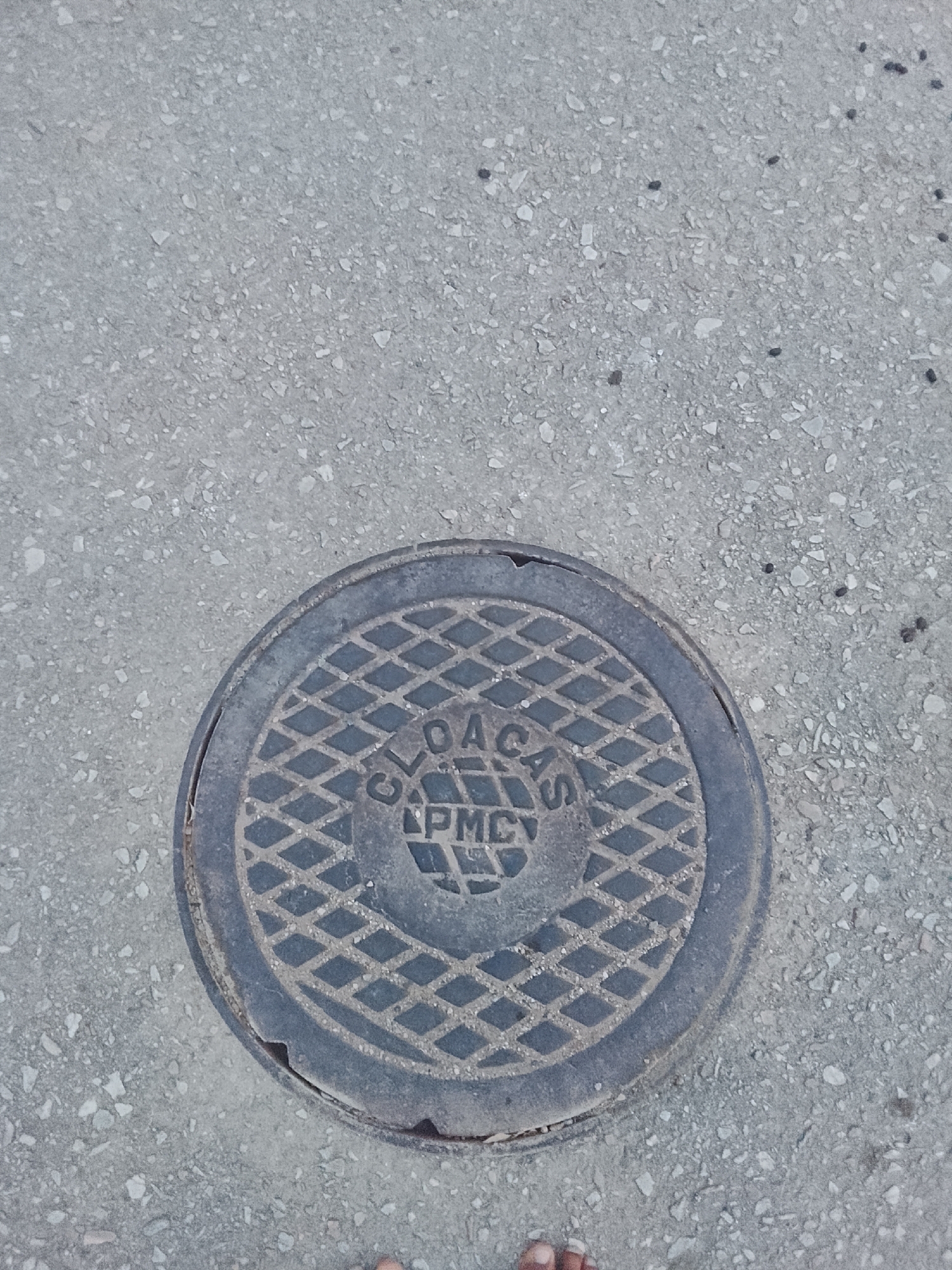 A low-angle shot depicts a circular manhole cover embedded in a paved surface. The cover is dark gray and features a grid-like pattern across its surface, with the text "CLOA CAS" and "PMC" inscribed in a central circular area. The pavement surrounding the manhole cover appears to be asphalt or concrete with a grainy texture. The scene is observed on a pedestrianized street, possibly in the city of Juan Griego, Venezuela, or any location where CLOA CAS and PMC have utilities. Partial view of foot/toes in the lower center of the frame.