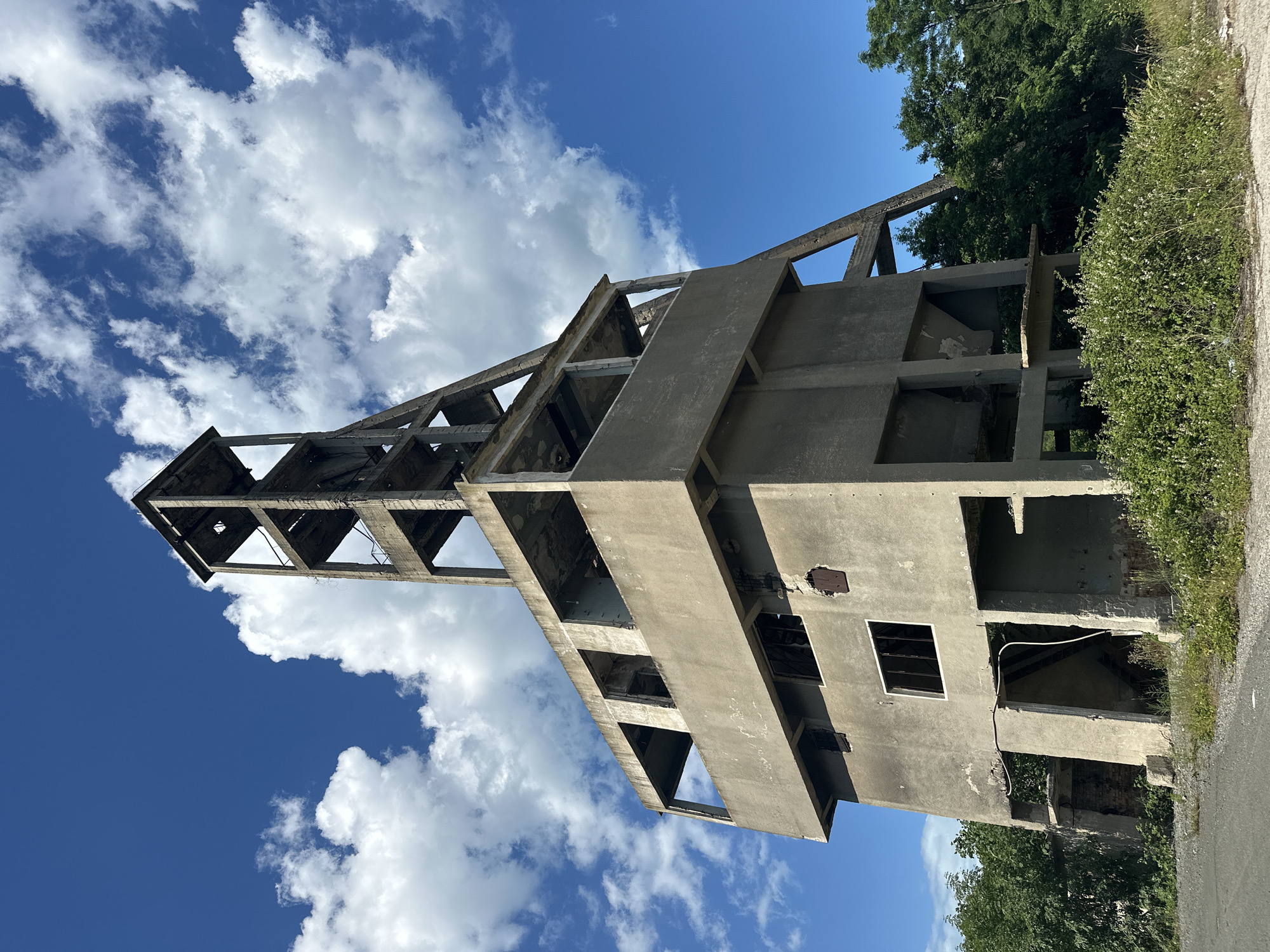 The image depicts an abandoned concrete industrial structure, likely a former mine shaft headframe, under a partly cloudy blue sky. The structure is tall and multi-leveled, with an open framework at the top supporting a concrete tower. The lower sections are more solid concrete, with visible openings that were once windows or doorways, many of which are now empty or damaged. The concrete appears weathered and stained, with some areas showing exposed rebar. Vegetation, including green bushes and white wildflowers, grows around the base of the structure and encroaches onto a gravel path in the foreground. Lush green trees are visible behind and to the right of the building. The overall impression is one of decay and nature reclaiming an industrial site. The location is identified as Rosen, Bulgaria. There are no people or animals visible in the image.