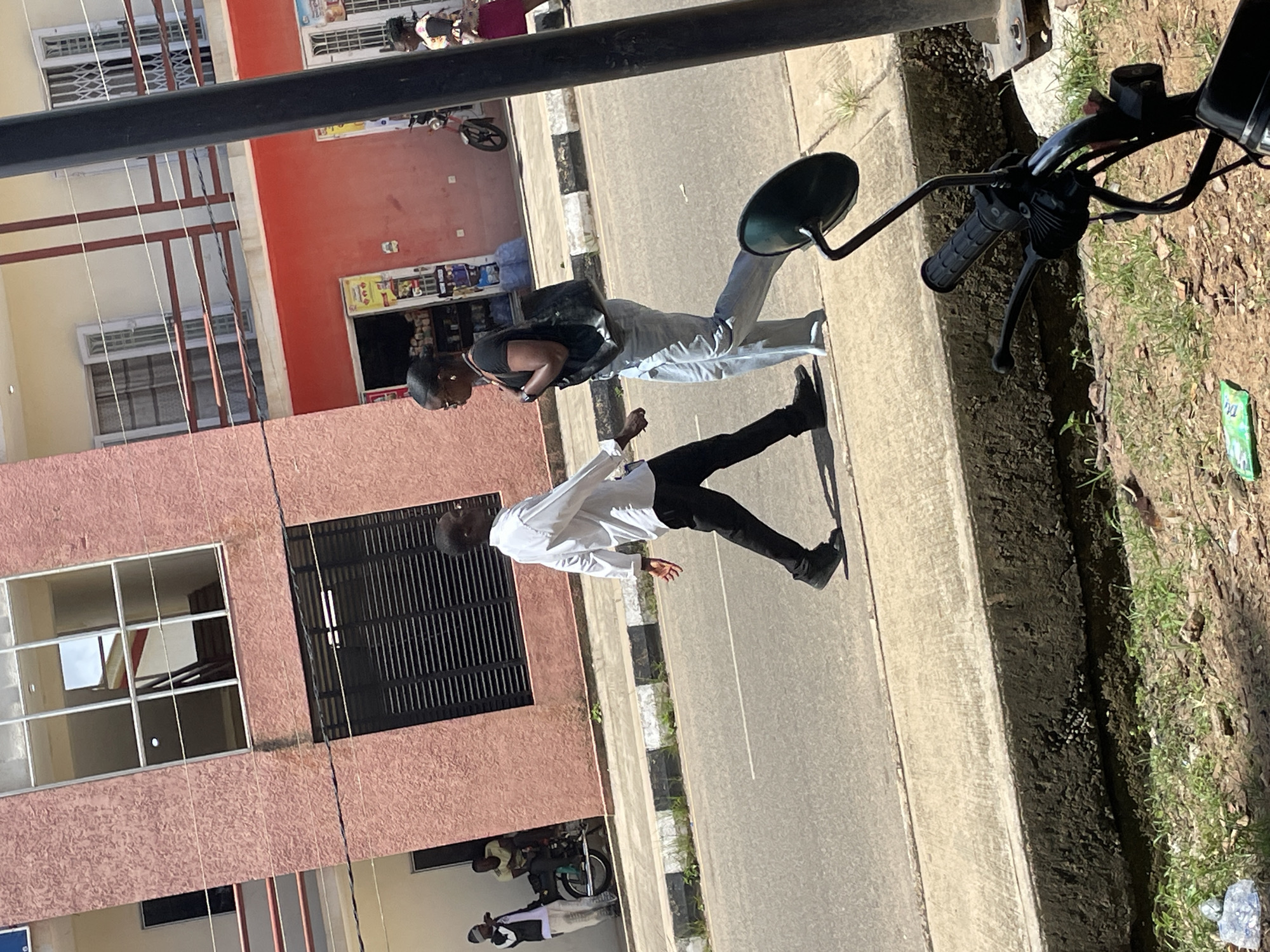 A man and a woman are walking on the street, with a building and a motorcycle in the background. The man, wearing a white long-sleeved shirt and black pants, is walking across the street. The woman, wearing a black t-shirt and light blue jeans, is walking on the sidewalk, carrying a black bag. In the background, a pink building with windows and balconies is visible. A motorcycle is parked on the side of the road, with two men standing near it. The scene appears to be a street in Jos, Nigeria, with typical urban elements like buildings, roads, and people going about their day.
