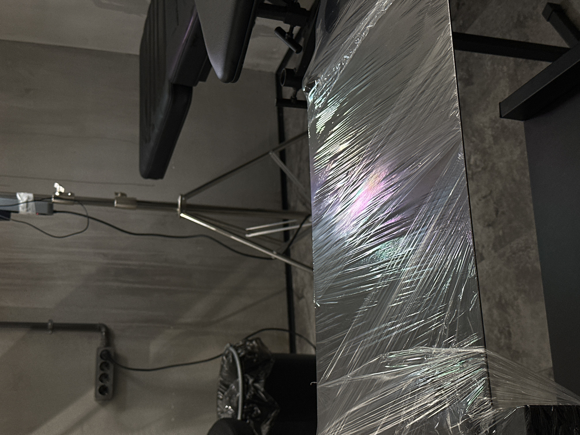 The image shows an indoor scene with various objects. On the right side of the image is a large object tightly wrapped in transparent plastic. Adjacent to it is a black structure or shelf attached to a gray wall. To the left, there's a metal stand with a dark, cushioned seat attached. The stand is positioned near a wall with an electrical outlet. The environment seems like a storage or studio space with utilitarian elements. The location, Moscow, Russia, is not evident from the image content.