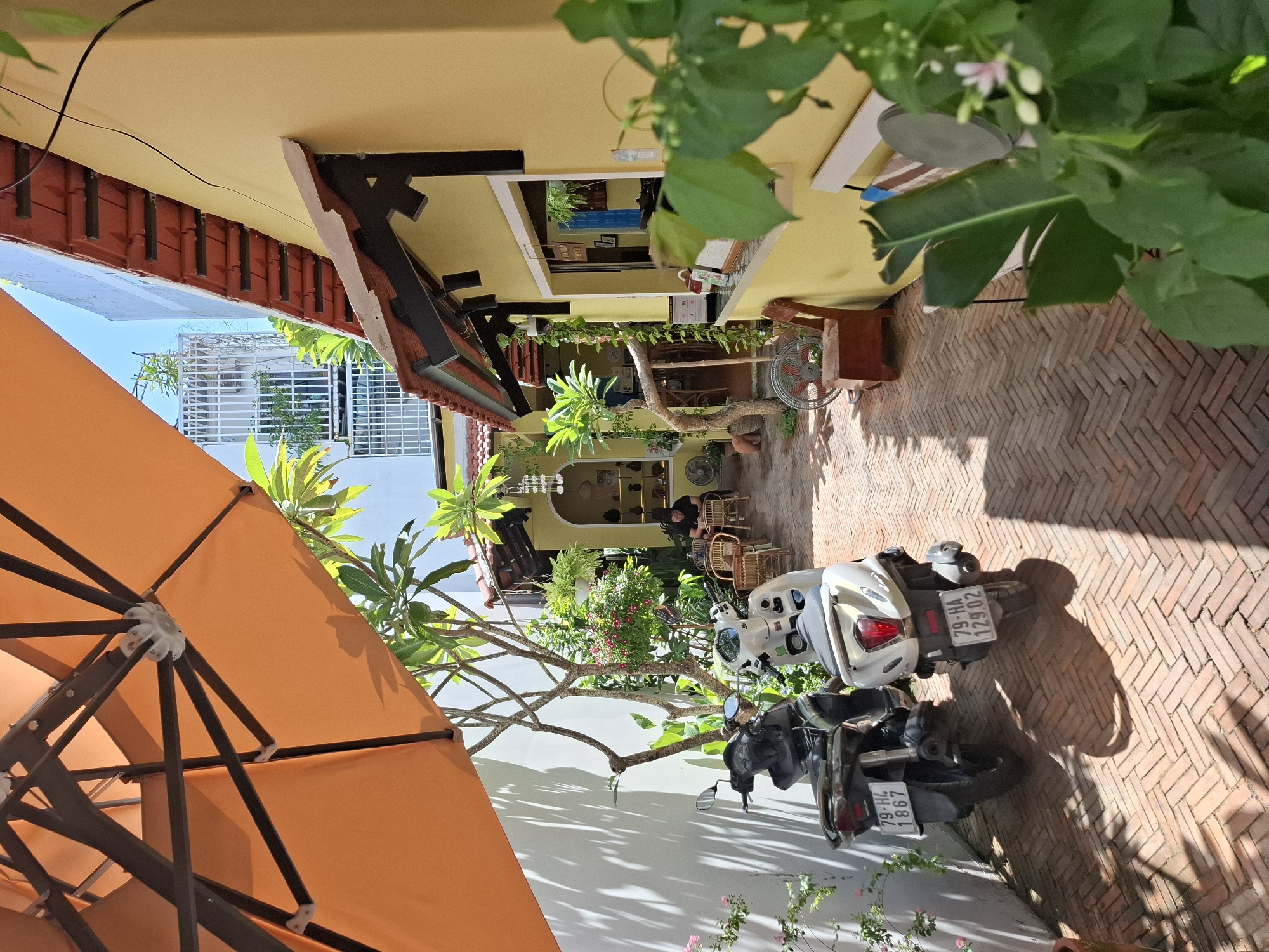 The image depicts an outdoor seating area of a cafe in Nha Trang, Vietnam. The foreground features two parked scooters, one black and one white, with license plates visible. An orange umbrella extends from the top left, casting shadows on the brick-paved ground and a white wall. Beyond the scooters, a person is seated at a table in the middle ground, surrounded by various plants and a few tables and chairs. The background reveals a yellow building with a tiled roof, a window serving as a counter, and in the distance, a modern white building with a fence. The scene suggests a casual, relaxed atmosphere, likely a cafe or restaurant in a city courtyard.