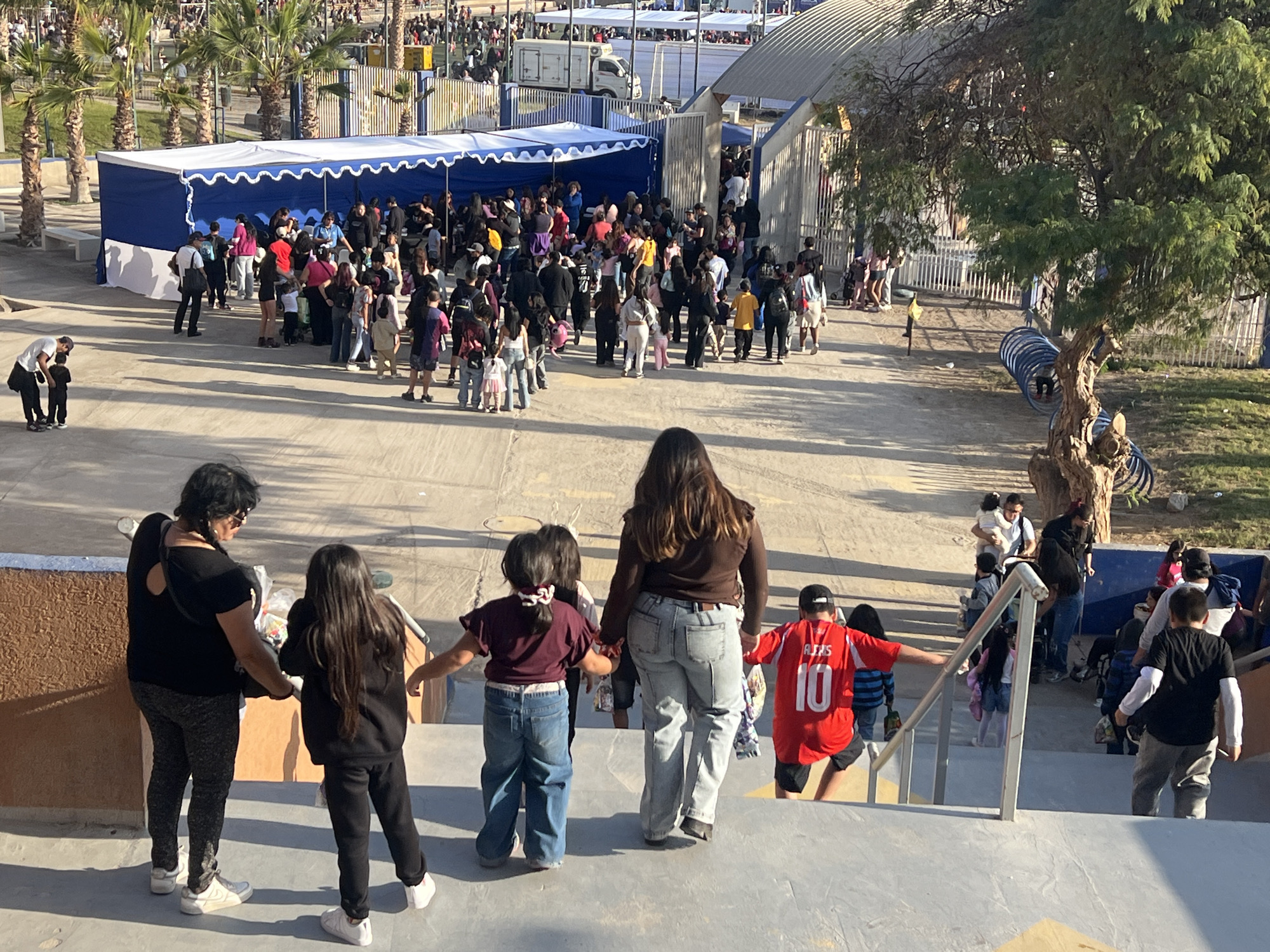 The image captures an outdoor scene likely in Arica, Chile, with a crowd of people gathered near a blue and white striped tent. Palm trees are visible in the background, suggesting a warm climate. In the foreground, a group of individuals, including adults and children, are walking on a paved surface and stairs. Some are dressed casually, with children in vibrant clothing. One child in the foreground wears a red jersey with the number "10". Further back, many people are seen milling around the tent, some appearing to be in line. The overall atmosphere suggests an event or gathering. The environment includes a paved walkway, stairs, a tent structure, fencing, and some greenery.