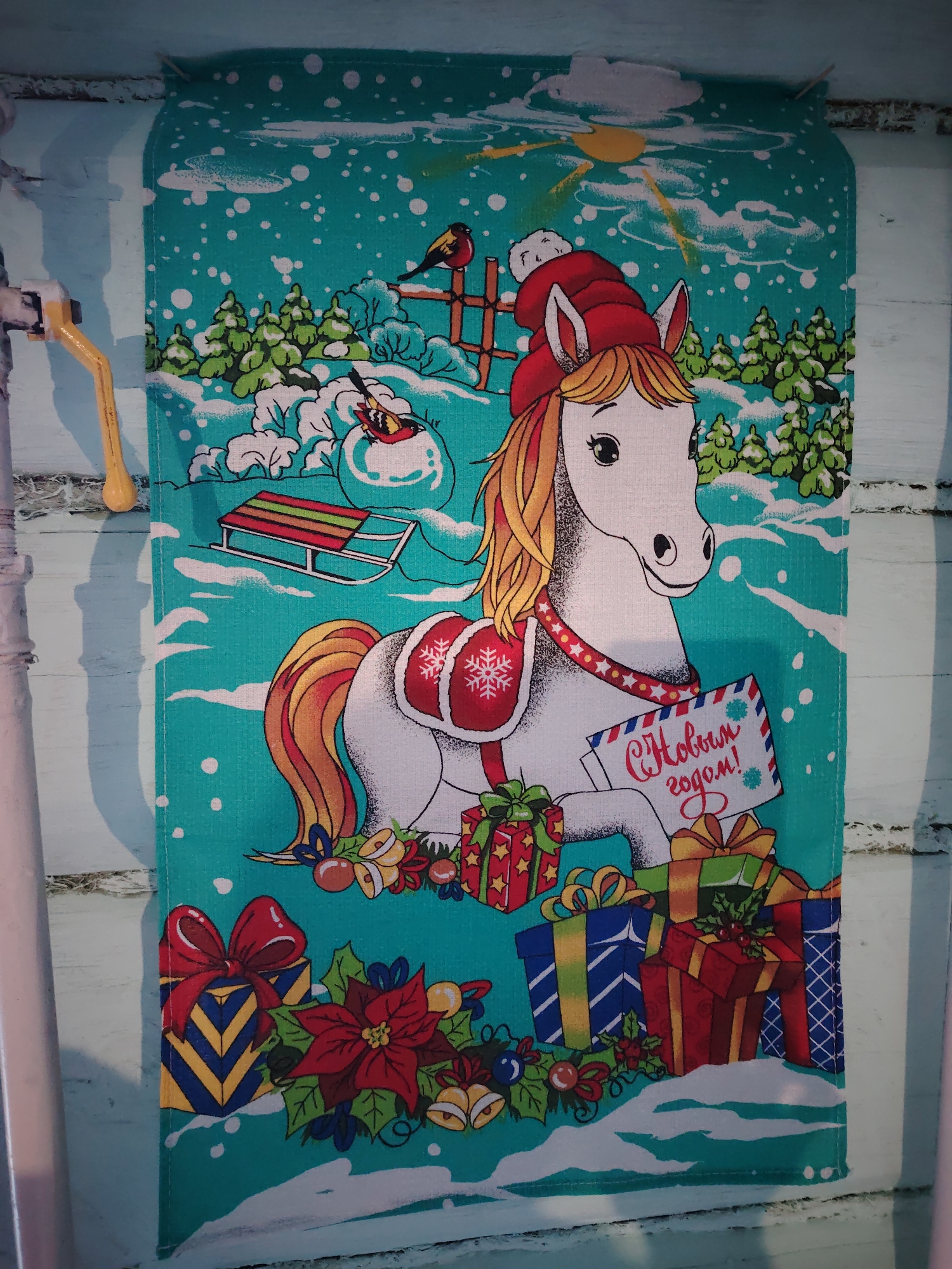 A decorative teal fabric piece, likely a towel or banner, depicts a cheerful winter holiday scene. The central subject is a stylized white horse with a golden mane and tail, wearing a red Santa hat and a red saddle adorned with a white snowflake. Around its neck is a red and white harness with stars. The horse holds an envelope with the Russian text "С Новым годом!" (Happy New Year!) in red.
The scene is set outdoors in a snowy landscape with evergreen trees, snowdrifts, and falling white snowflakes. In the background, to the left of the horse, a snowman wears a red cap and is accompanied by a small red-breasted bird perched on a wooden fence. Another small bird is visible near the snowman. A red and green striped sled rests in the snow. A bright yellow sun peeks from behind clouds in the upper right corner.
Numerous festively wrapped gift boxes with colorful bows, poinsettias, and decorative bells, holly, and berries are arranged around the base of the horse, in the foreground. The overall atmosphere is joyful and celebratory, indicative of the New Year or Christmas holidays. The fabric is hung against a light blue wooden wall, with a yellow spigot or handle visible on the left side of the frame. This media file was taken in Сусады-Эбалак, Russia.