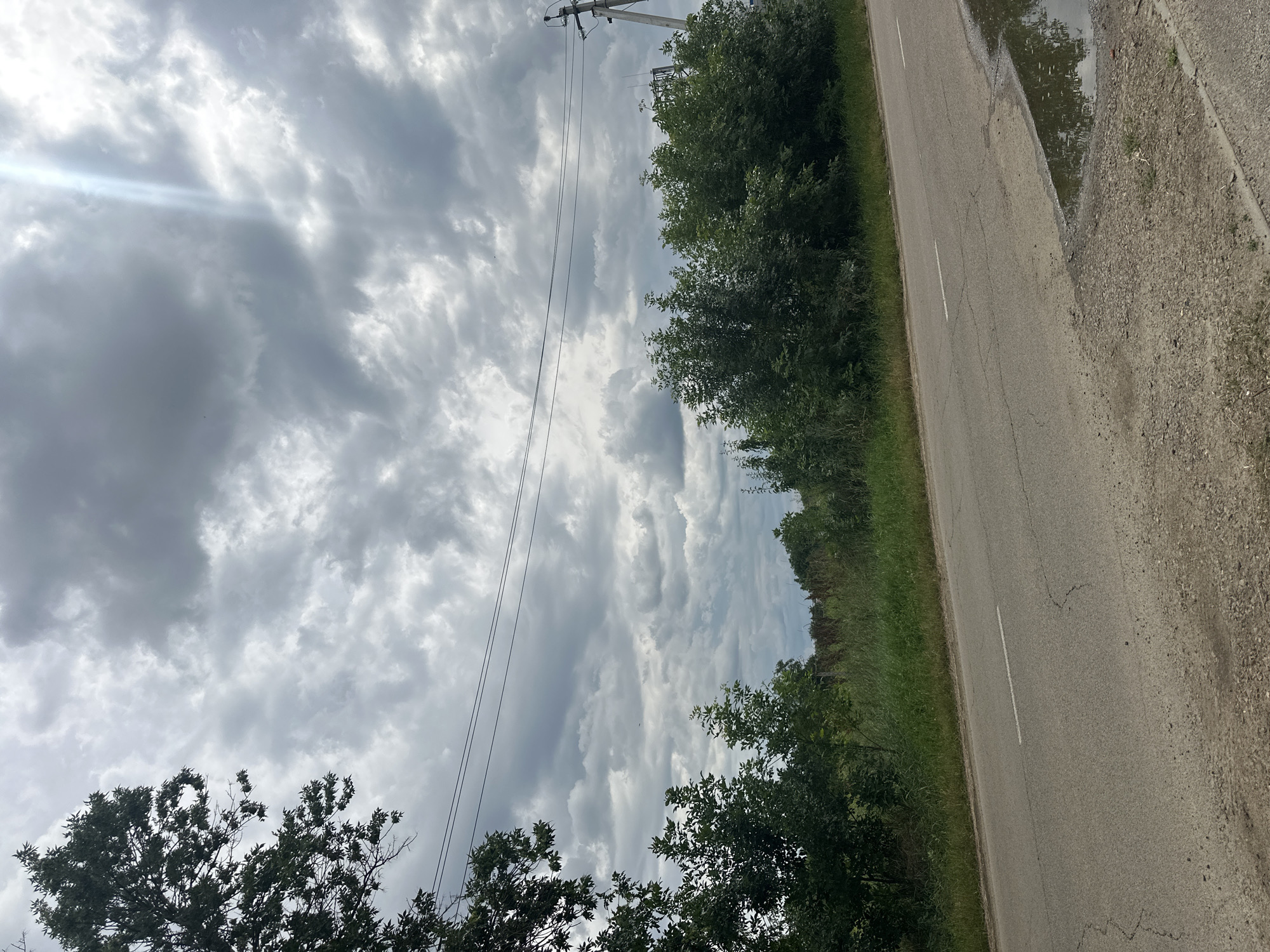 An asphalt road stretches across the lower portion of the frame, a white line marking its center. A puddle reflects the overcast sky on the right side of the road. On the left, lush green trees and vegetation border the road, with taller trees framing the left edge of the image. In the background, a dense line of trees creates a dark green horizon. Above, a dramatic sky is filled with heavy, grey clouds, broken by a bright shaft of sunlight piercing through from the upper right. Power lines are strung across the sky. The scene appears to be a rural road in Tbilisskaya, Russia, under cloudy weather conditions.