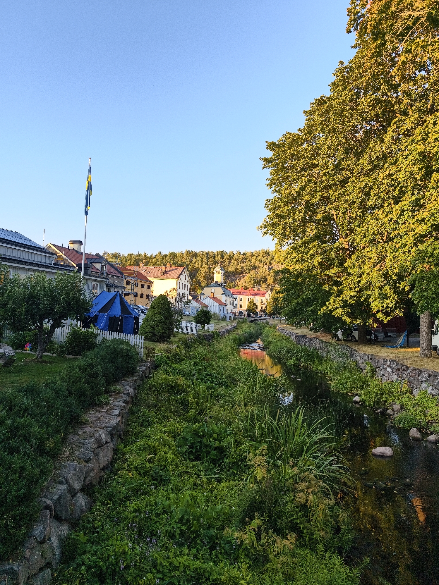 This is a wide-angle, eye-level, outdoor photograph taken in the city of Söderköping, Sweden, at golden hour. The scene depicts a small town along a narrow river. On the left bank of the river, houses with red-tiled roofs are visible, along with a blue and black tent pitched in a grassy area. A flagpole with the Swedish flag is prominent in the foreground. The right bank of the river is lined with large, lush green trees, their leaves illuminated by the setting sun. The river itself is calm and reflects the surrounding environment. The overall impression is one of a peaceful, idyllic summer evening in a Swedish town.