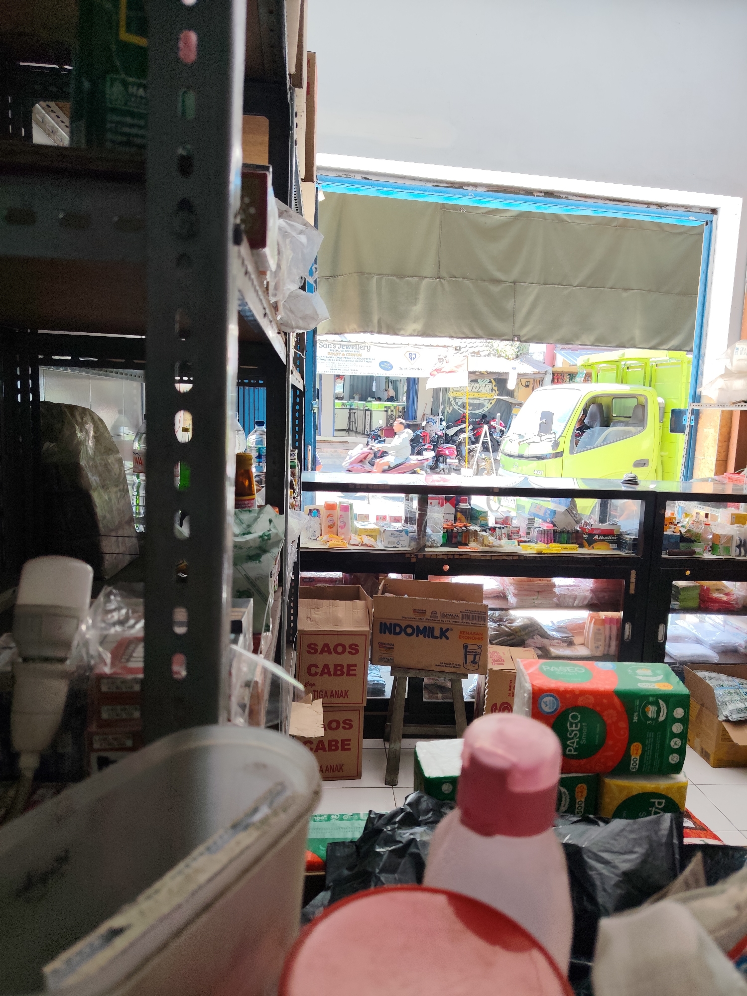 The image shows the interior of a small convenience store in Dampit, Indonesia, looking out towards a street scene. In the foreground, shelves are filled with various products, including boxes of "SAOS CABE" (chili sauce) and "INDOMILK" (milk). A green truck is visible through an open doorway, along with parked motorcycles and people on the street. The store appears to be well-stocked with consumer goods, and the overall impression is of a typical neighborhood shop.