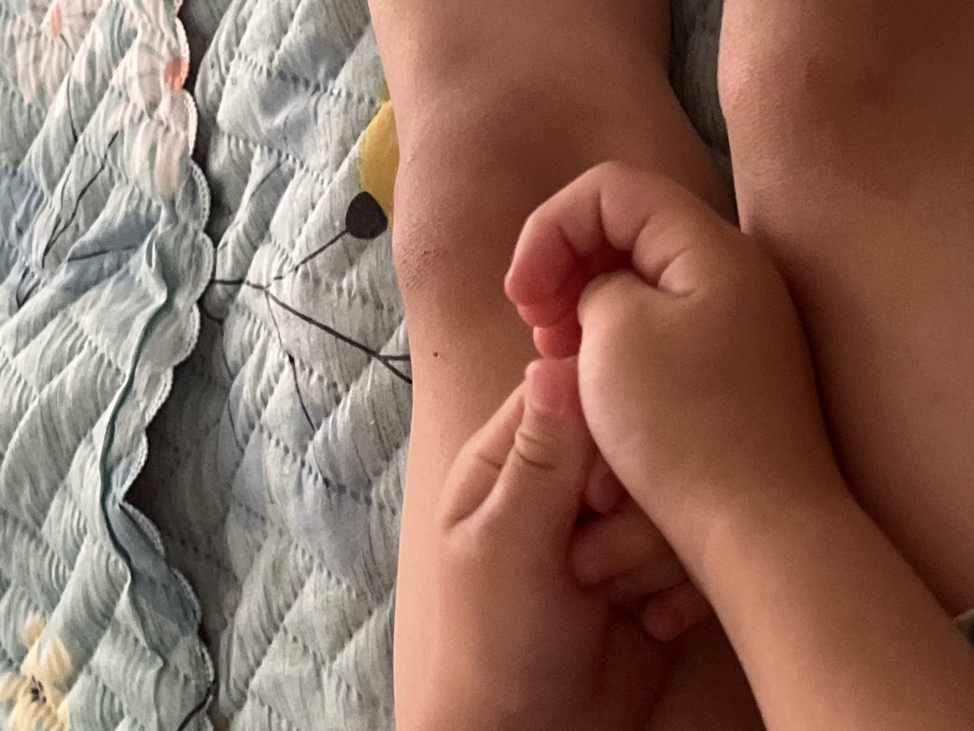 The image shows a close-up view of two children's hands interlaced against a backdrop of a quilted blanket. The blanket has a light blue and green floral pattern with scalloped edges. One child's hand is positioned in front, with fingers curled inward, and the other child's hand is beneath it, also with fingers curled. The children's skin is light-toned. The scene appears to be indoors, possibly on a bed or couch. There is no discernible indication of the city of Astana, Kazakhstan, or any urban environment.
