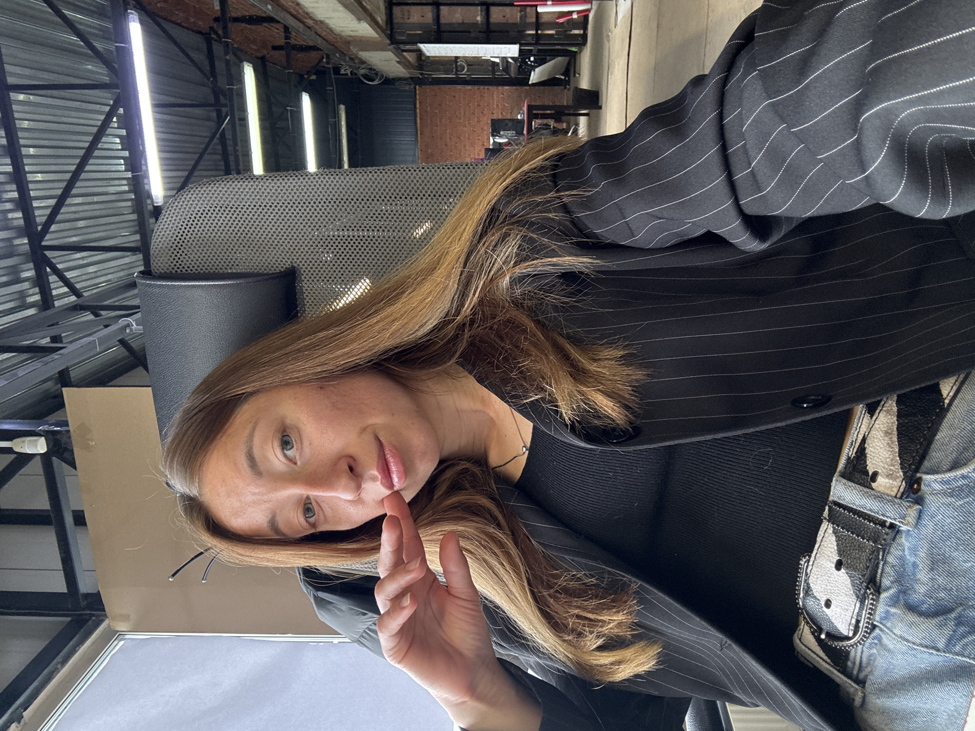 A woman is seated in an office chair in Chaikovsky, Russia. She is wearing a black pinstripe blazer over a black top and blue jeans with a patterned belt. The office setting has exposed metal beams and fluorescent lighting, suggesting an industrial or renovated space. The background shows a glimpse of a brick wall in the distance. The woman is looking at the camera with a slight smile, and her hand is raised with her fingers extended.