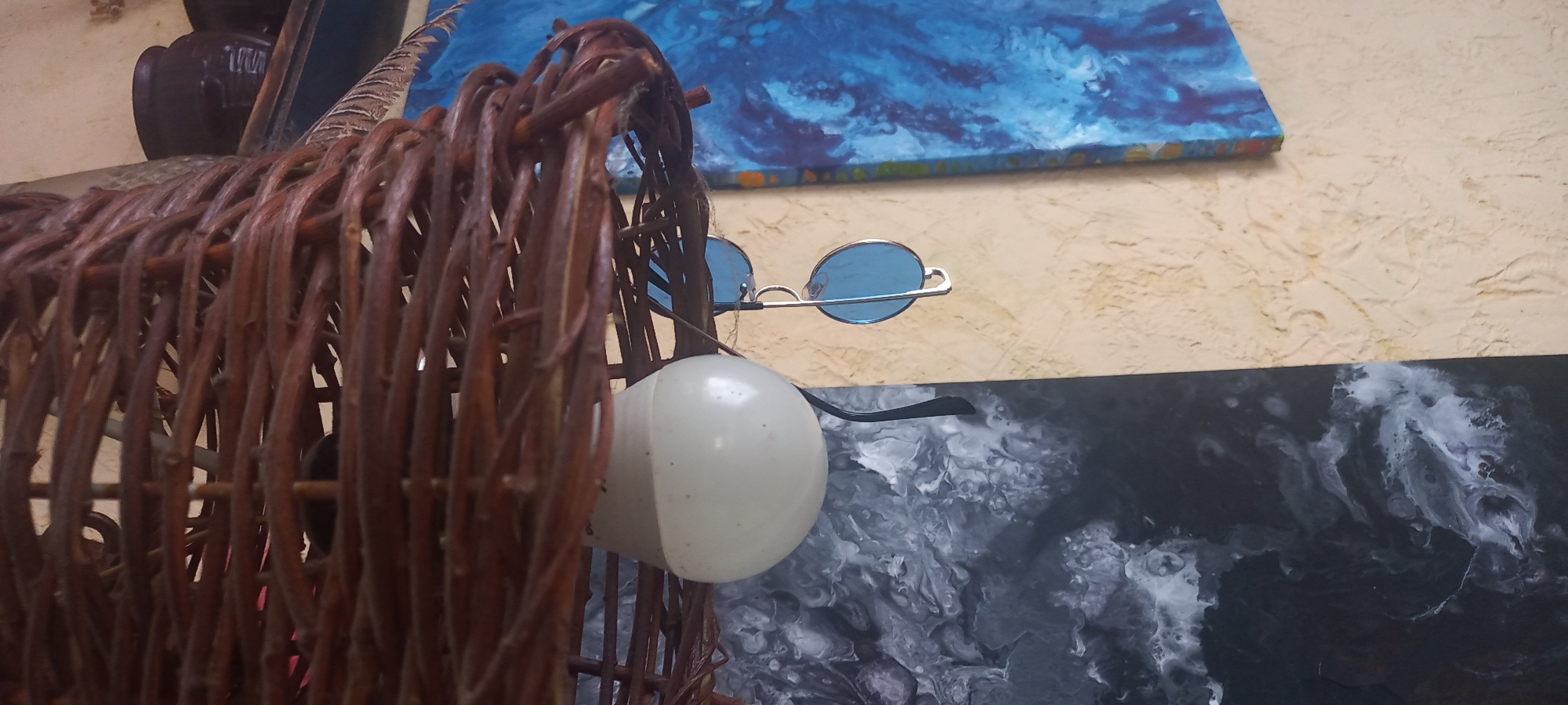 The image shows a close-up of various objects arranged against a textured wall. A woven brown wooden structure, possibly a basket or animal figurine, occupies the left side of the frame. A white light bulb and blue-tinted round sunglasses are situated near the wooden structure. Two paintings hang on the wall: one with a blue and white abstract design and another with a black and white marble-like pattern. The scene is generally cluttered with diverse items. The location, Kryvyi Rih, Ukraine, is not inherently evident from the image itself.