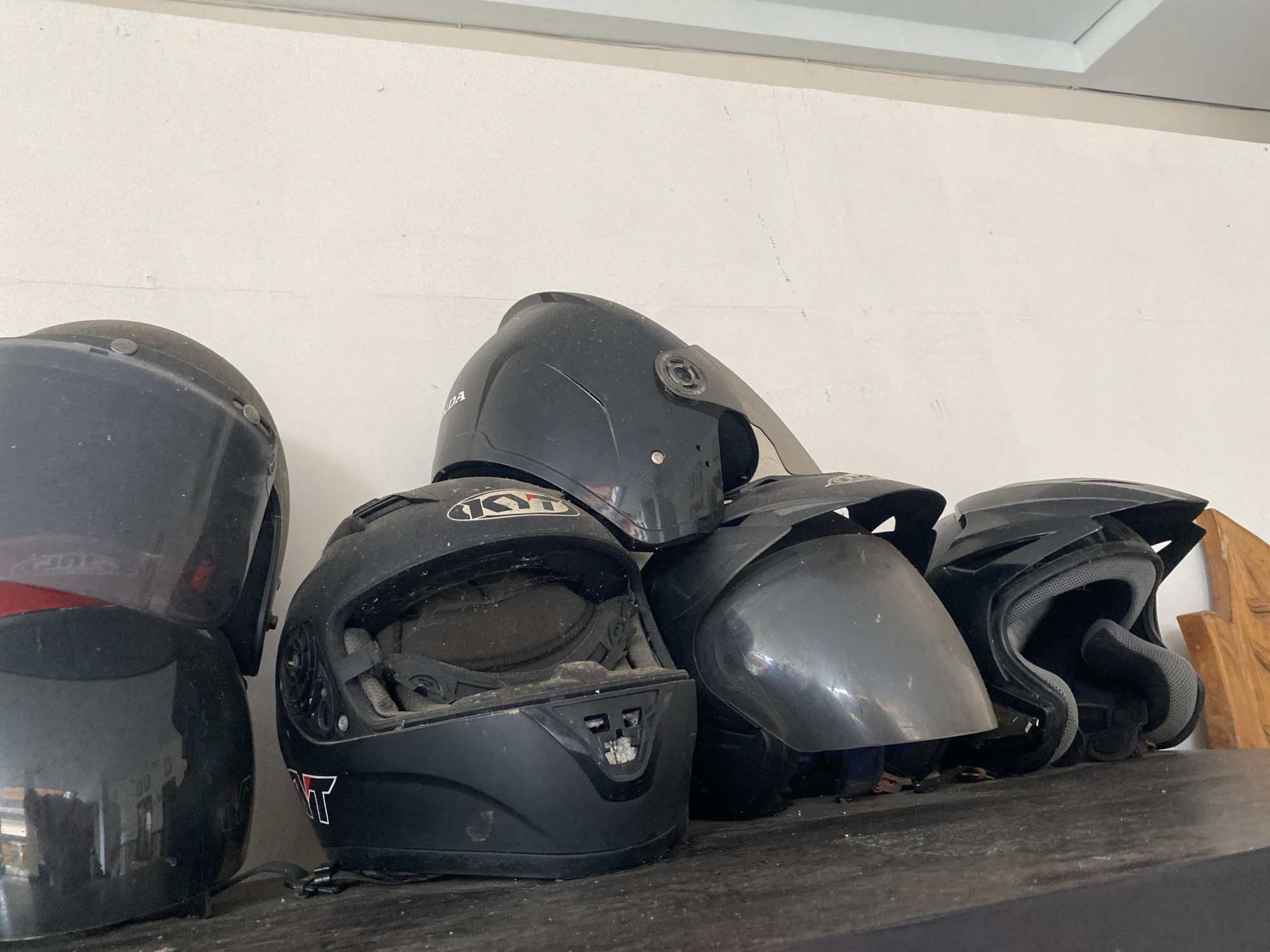 A low-angle, medium shot depicts a collection of five black motorcycle helmets arranged on a dark, dusty surface against a white wall. The helmets are of varying styles and conditions, appearing worn and dusty. A "KYT" brand helmet is visible. The background is a plain white wall with a slight texture. The lighting is soft and diffused. The location is Nusa Dua, Indonesia.