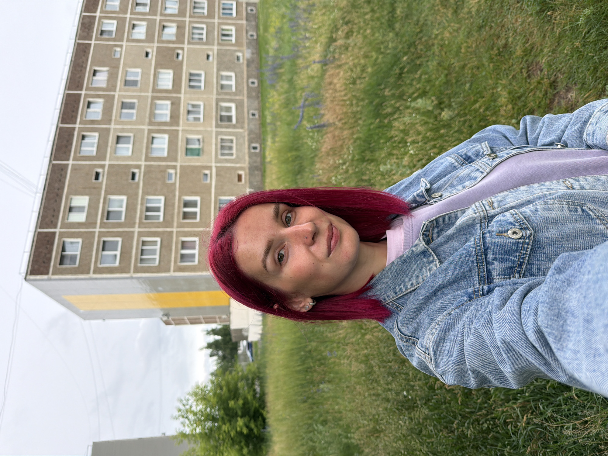 In the city of Chaikovsky, Russia, a young woman with vibrant pink hair stands in front of a tall, multi-story apartment building. The building, constructed from beige concrete with brown trim, features numerous rectangular windows and balconies. A striking vertical band of bright yellow paint runs along one side of the building. The woman is wearing a light blue denim jacket over a lavender sweatshirt and a silver earring in her left ear. She has a subtle nose piercing. The foreground is filled with lush, green grass, with some areas showing sparse purple flowers. The sky above is overcast and grey.