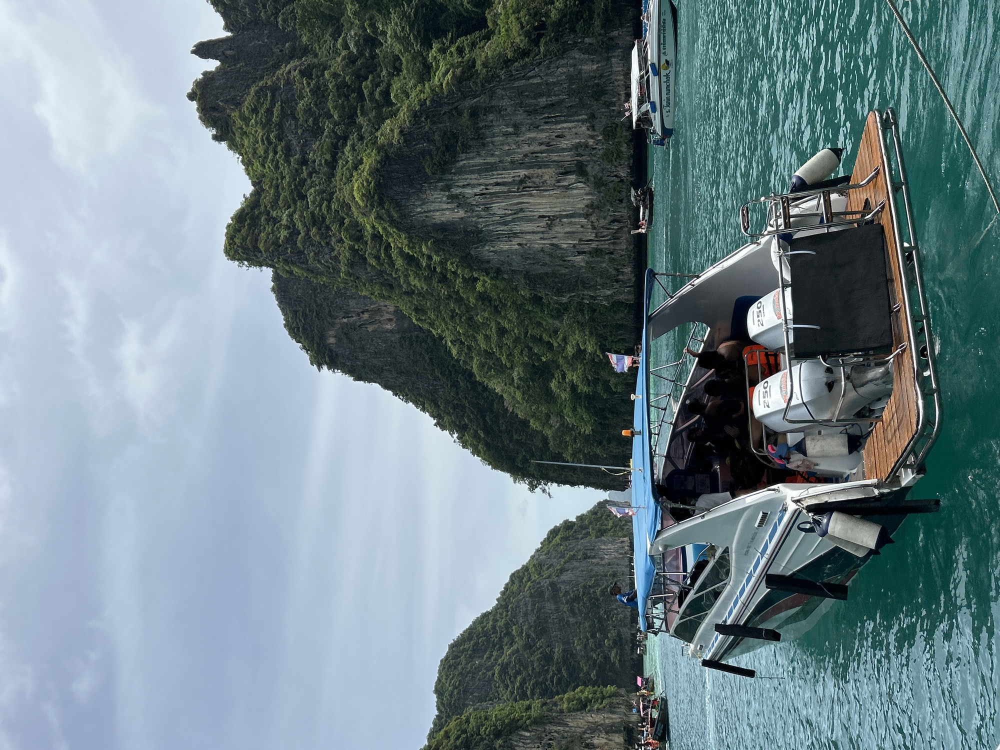 A motorboat with a blue canopy is anchored in turquoise water, surrounded by lush green limestone cliffs under a cloudy sky. Several people are visible on the boat, some sitting under the canopy and others standing. The boat has two outboard motors labeled "250." In the background, other boats, including a white speedboat with text in Thai, are scattered across the water. The scene depicts a scenic location, likely in the waters off Ao Nang, Thailand, known for its dramatic karst landscapes and popular boat tours.