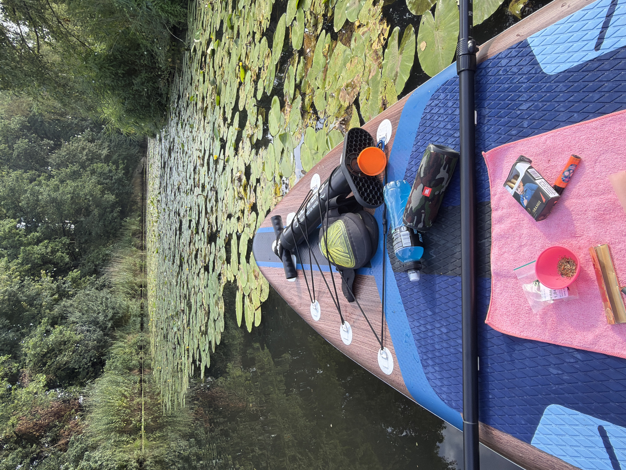 The image shows a paddleboard floating on a canal in Boxtel, Netherlands, with numerous lily pads covering the surface of the water. On the paddleboard, there is a black pump with an orange cap, a blue water bottle, a camouflage-patterned speaker, a pink towel, a small pink bowl containing dried herbs, a packet of cigarettes, and what appears to be a vaping device. The paddleboard is positioned with the front facing forward, indicating it is either stationary or moving downstream. The surrounding environment consists of lush green trees and vegetation lining the canal banks, suggesting a natural and serene setting. The overall scene depicts a recreational activity taking place on a calm waterway.