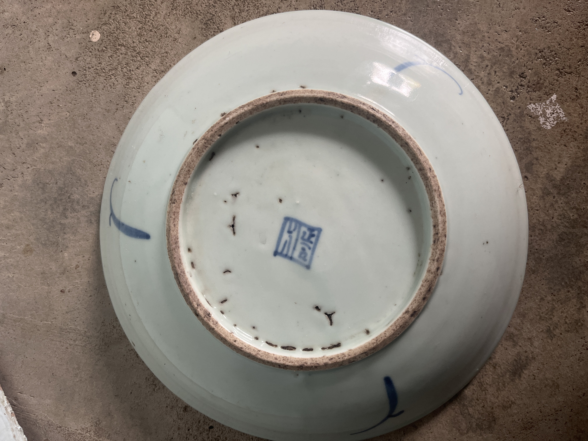 The image shows the underside of a light blue ceramic plate resting on a textured concrete surface. The plate has a raised, unglazed rim. Faint blue brushstrokes, likely decorative elements, are visible on the outer surface of the plate. In the center of the underside, there is a blue square stamp with what appear to be Chinese characters inside. The concrete surface is uneven and shows some discoloration and debris. There are no people, animals, or signs of the city of Meliau Hulu, Indonesia. The scene is a close-up, overhead shot of the plate and its immediate surroundings.