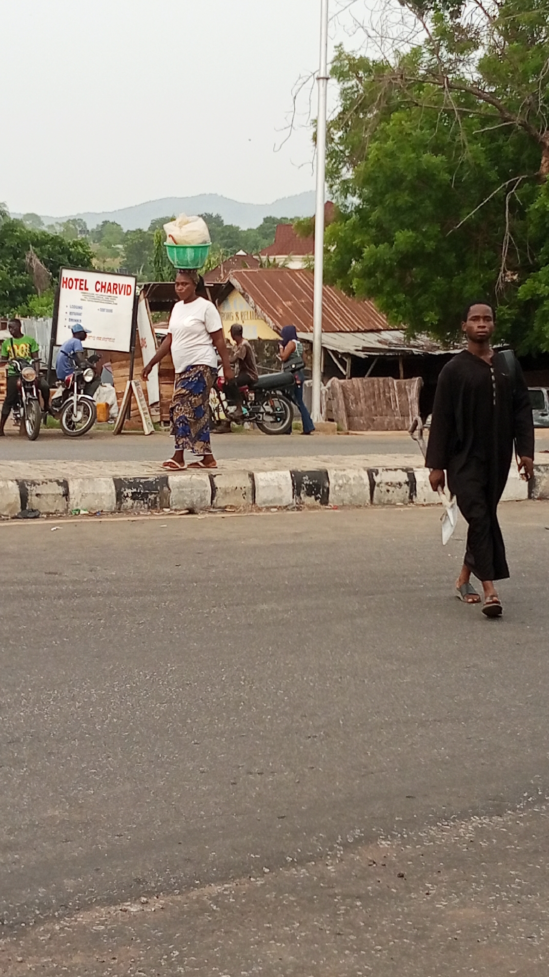 In Adankolo, Nigeria, a street scene unfolds on a grey asphalt road, bisected by a concrete curb painted with black and white markings. A woman walks along the curb carrying a white bag and green basin on her head. She wears a white t-shirt and printed blue pants. To her right, behind the curb, men are on motorcycles near a sign that reads "HOTEL CHARVID." One man is walking along the curb to the right of the woman with an object in his hand, he wears a long black tunic and sandals. A utility pole is visible near the sign. Further behind, buildings with corrugated metal roofs, trees and distant hills contribute to the background.