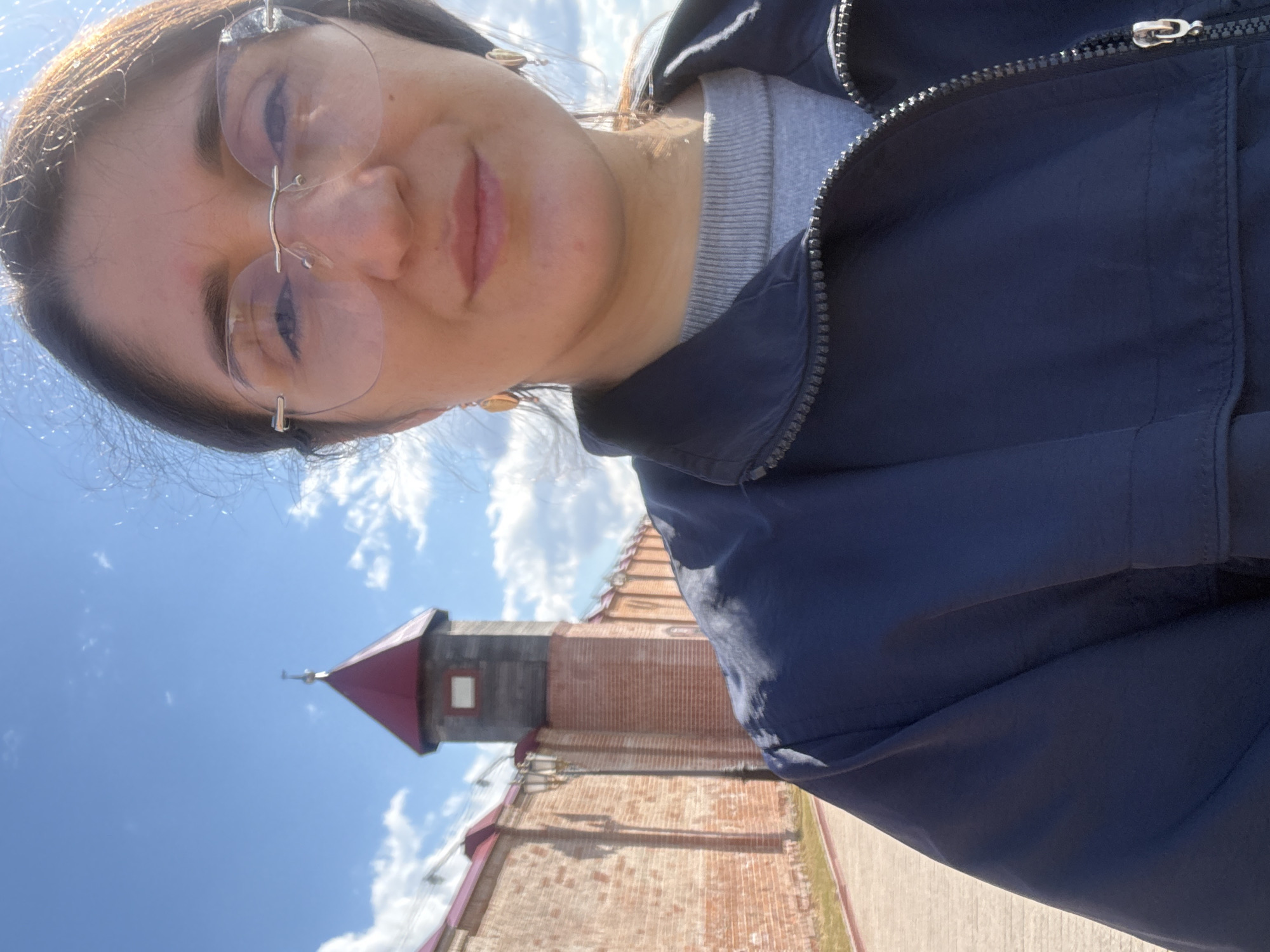 A woman is taking a selfie in front of the Tobolsk Kremlin in Tobolsk, Russia. The woman has dark hair and is wearing glasses, a gray sweater, and a dark blue jacket. The background shows a partially-visible red-brick building with a distinctive tower, a light blue sky, and white clouds.