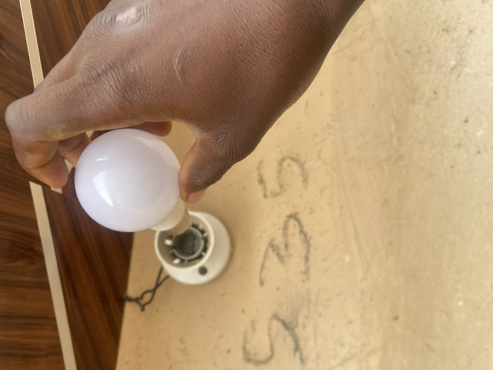 A person's dark-skinned hand is holding a white LED light bulb near a light socket. The light bulb is spherical and emits a bright white light. The light socket is white and appears to be mounted on a textured, light brown wall. In the background, there is a wooden surface with a horizontal white stripe. The wall also has faint markings that resemble numbers or symbols. The image appears to be taken outdoors, possibly in a residential or commercial area in DU, Nigeria.