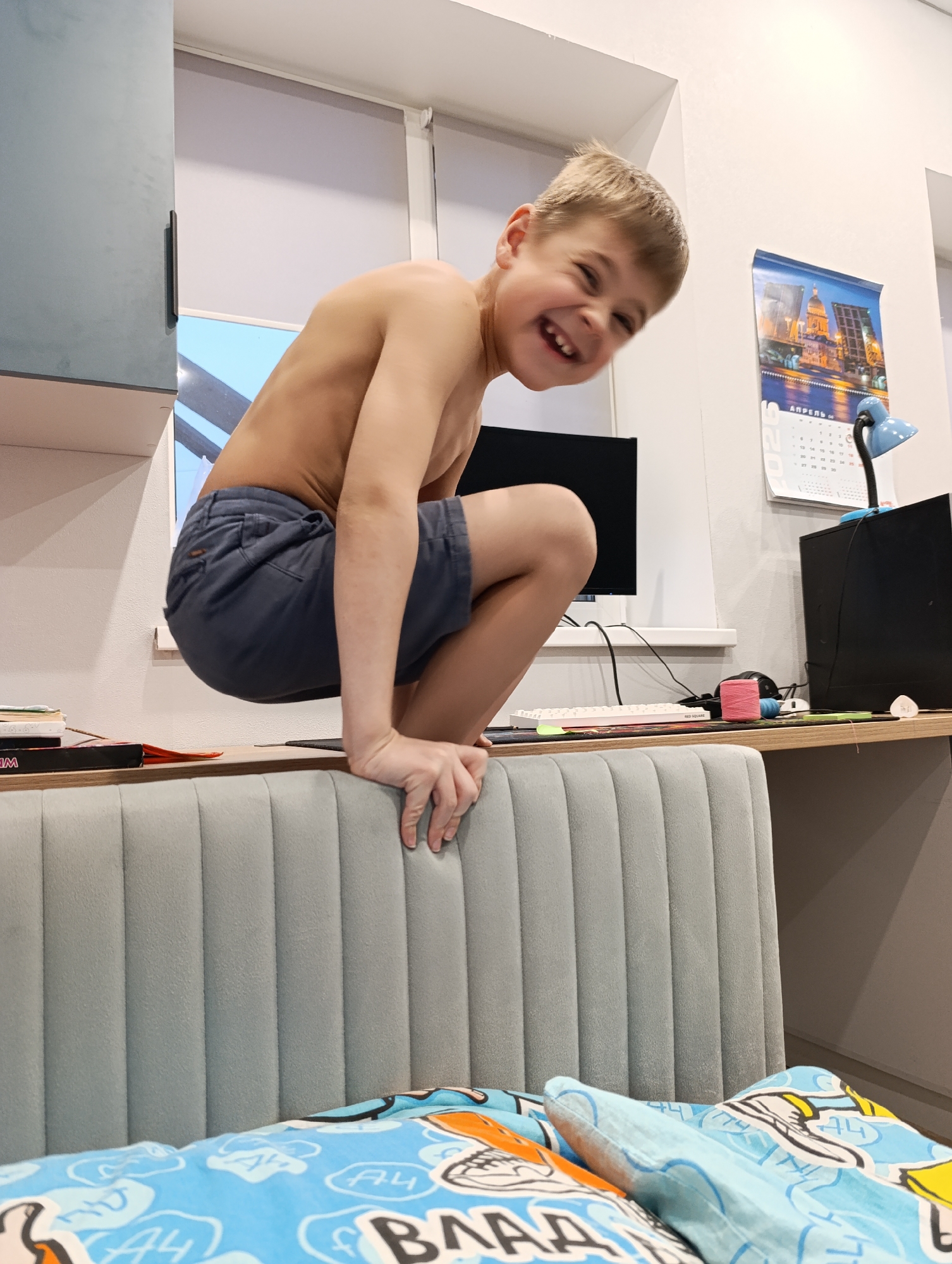 A fair-skinned child squats on the edge of a light blue headboard of a bed in a room in Chaikovsky, Russia. The child is shirtless, wearing dark blue shorts, and smiles with visible teeth. Behind the child, a desk is visible with a computer monitor, a white keyboard, a small blue desk lamp, and a black computer tower. A calendar hangs on the wall above the desk. The bed has patterned blue and orange bedding. A blue cabinet is visible on the left side of the frame.