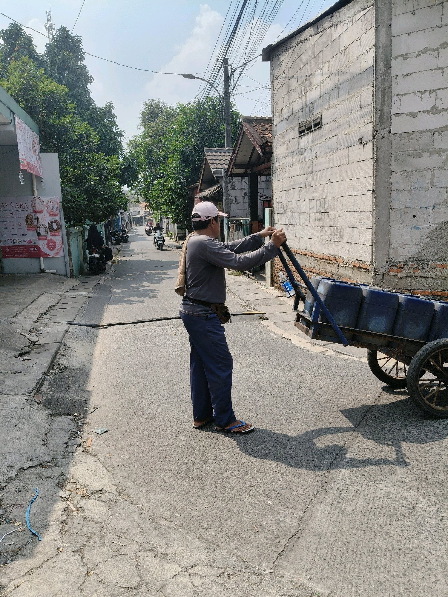 A man wearing a gray long-sleeved shirt, blue pants, a white cap, and flip-flops is pushing a cart with blue barrels down a paved street in a residential area. The cart has two wheels and a wooden frame. The man is holding the handles of the cart with both hands and appears to be looking forward. A tan towel is draped over his left shoulder. A brown pouch is attached to his belt. The street is lined with buildings on both sides, including a building constructed of white cinder blocks with visible mortar lines. There are trees and utility poles with many wires visible in the background. The sky is partly cloudy. The scene is illuminated by daylight. The location is identified as South Tangerang, Indonesia based on the provided context.