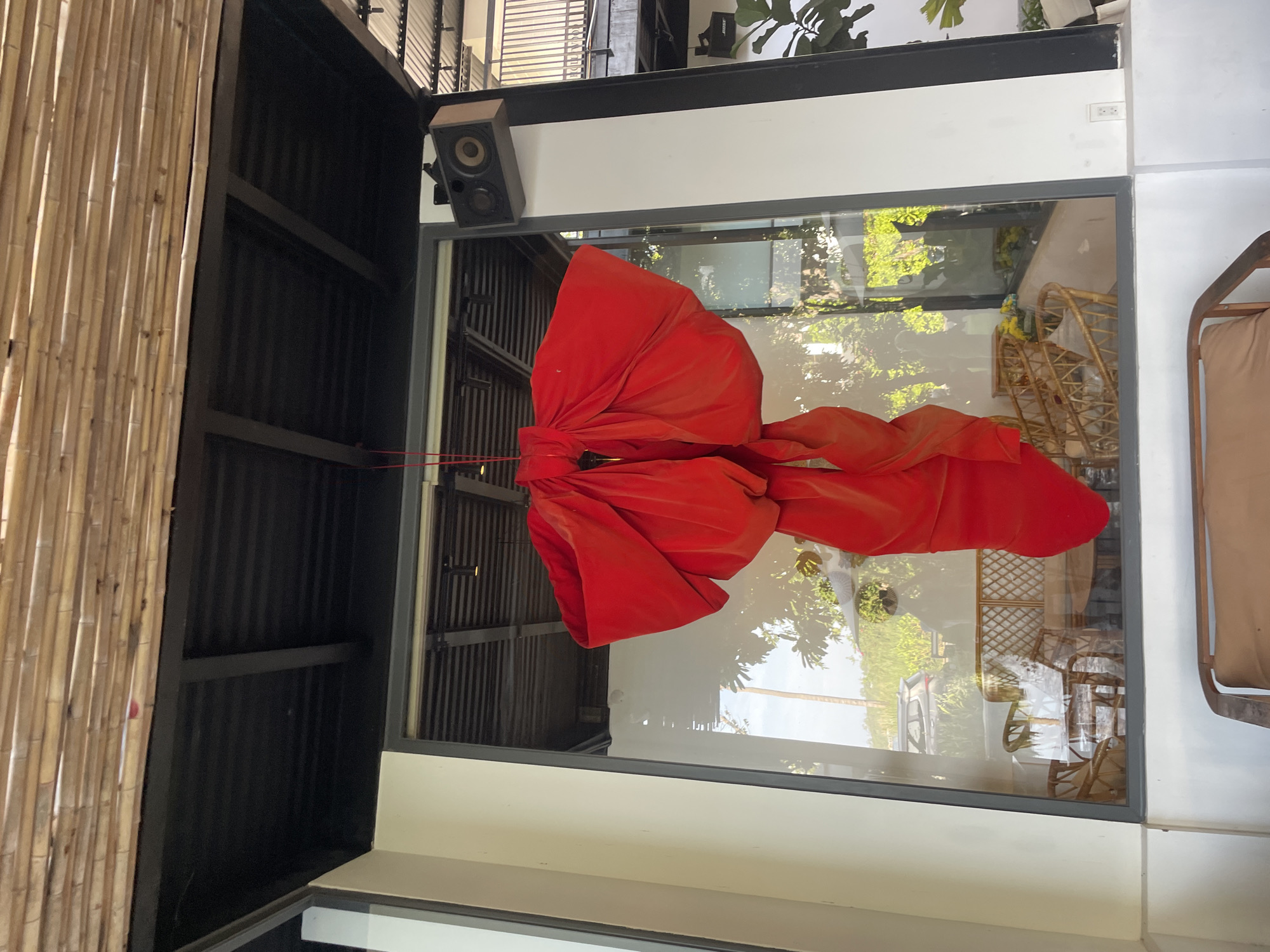 The image shows a large red bow hanging from a ceiling over a window. The bow is made of velvet material and appears to be a decorative item. Inside the window, there is a reflection of furniture that appears to be wicker chairs and a table. The background outside the window is blurred, but it seems to be an outdoor setting with greenery. There is a speaker mounted on the wall to the right of the window. The ceiling above is made of bamboo. The scene is likely an outdoor or semi-outdoor space, possibly a cafe or a restaurant, in Nam Nha Trang Ward, Vietnam. There are no people visible in the image.