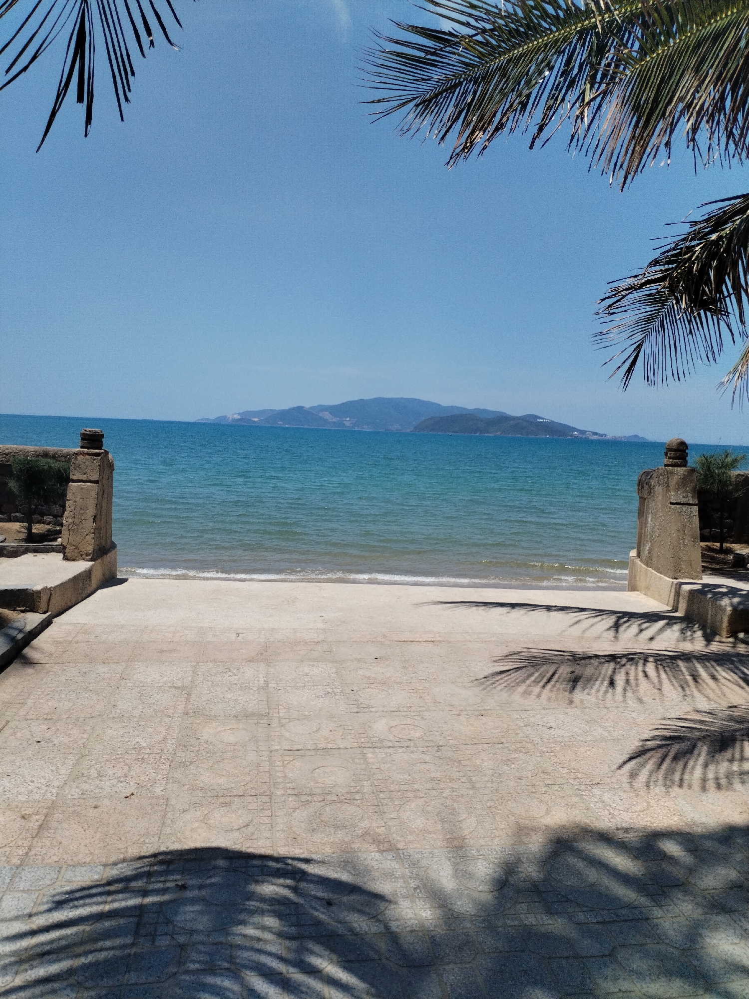 View of the beach and ocean from a stone pathway in Nha Trang Ward, Vietnam. The pathway is bordered by low stone walls or platforms on either side. Palm trees overhang the pathway, casting shadows on the stone tiles. Beyond the beach, there is a body of water with a mountainous landmass in the distance under a clear, blue sky.