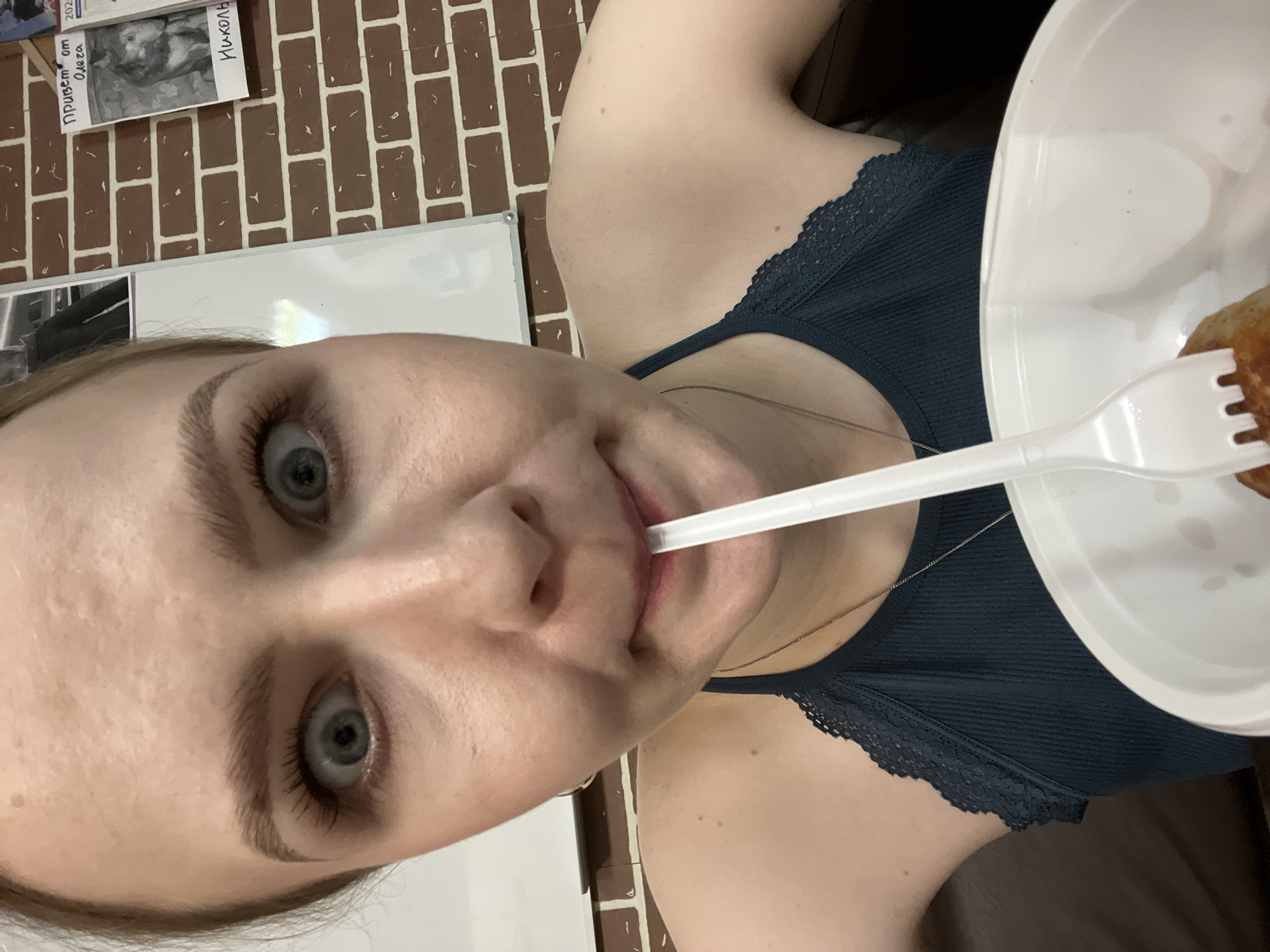 A young woman is centered in a selfie-style, close-up shot. Her face is the dominant feature, with wide eyes looking directly at the camera. A white plastic fork is held in her mouth. She is wearing a dark blue top with a lace trim. A white disposable plate is partially visible, containing some food. The background includes a wall with a brick pattern and a whiteboard with some notes and photos attached. The whiteboard contains what appears to be a photo of the painting "Ivan the Terrible and His Son Ivan" by Ilya Repin, with a note saying "Hello from Oleg to Nikola". The picture may be taken in Khimki, Russia.