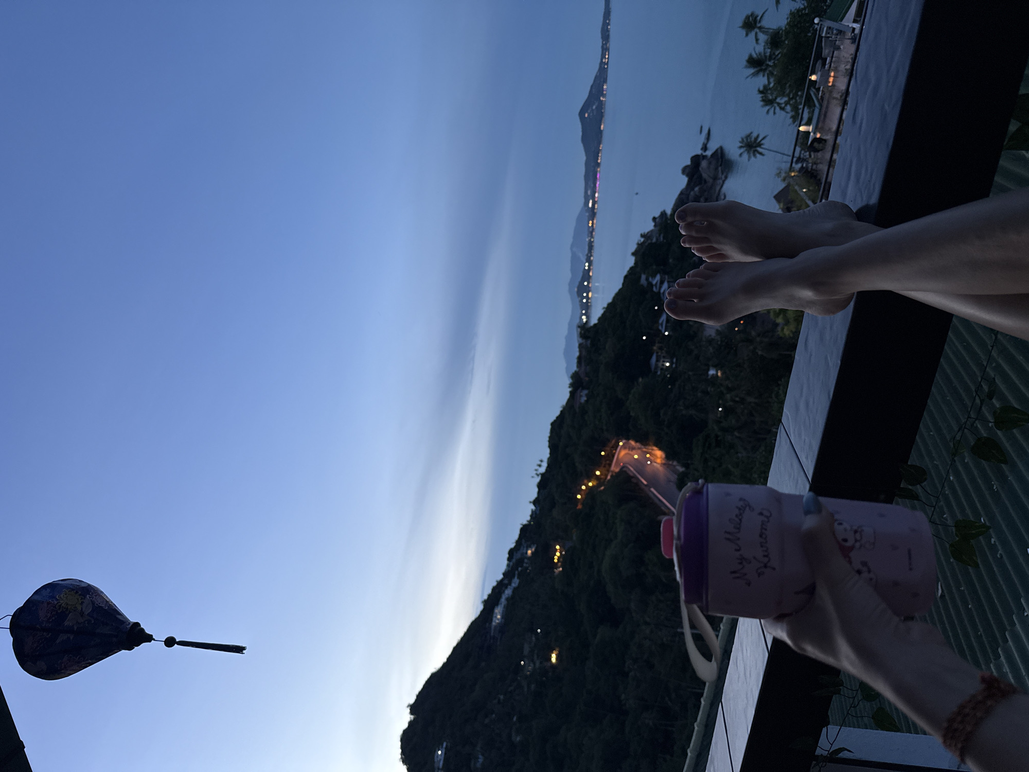 The image shows a first-person perspective of a person holding a pink water bottle with "My Melody Kuromi" written on it. Their bare feet are resting on a dark railing, with their legs crossed. The background features a view of a coastal road winding up a lush green, mountainous landscape. In the distance, the ocean meets the horizon under a twilight sky. Lights from buildings are scattered across the hillside and along the coast. A decorative lantern hangs in the upper left corner. The location appears to be a vantage point in Ko Samui, Thailand, likely from a balcony or terrace overlooking the sea.