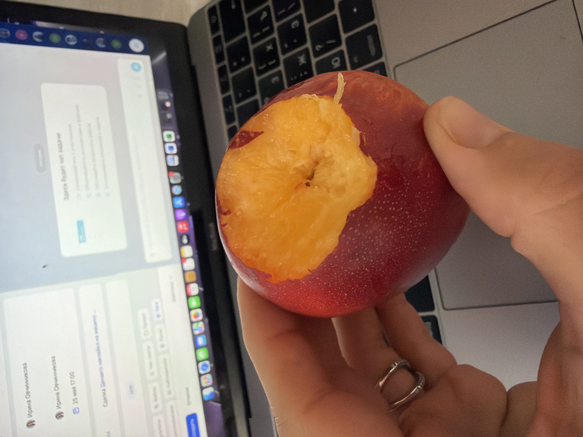 A person is holding a partially eaten nectarine in their hand, with a laptop in the background. The screen of the laptop shows a chat interface with messages in Russian. The person is wearing a ring on their finger. The scene appears to be indoors, possibly at a desk, with a keyboard visible in the background. Based on the OCR text, the chat may be related to "Ирина Овчинникова" and a "Deal Denieto" involving stickers for a car. The date "25 мая 17:00" is also mentioned, suggesting the time of the message. The location in the city of Chaikovsky, Russia, is not directly visible in the image and cannot be determined from the available information.