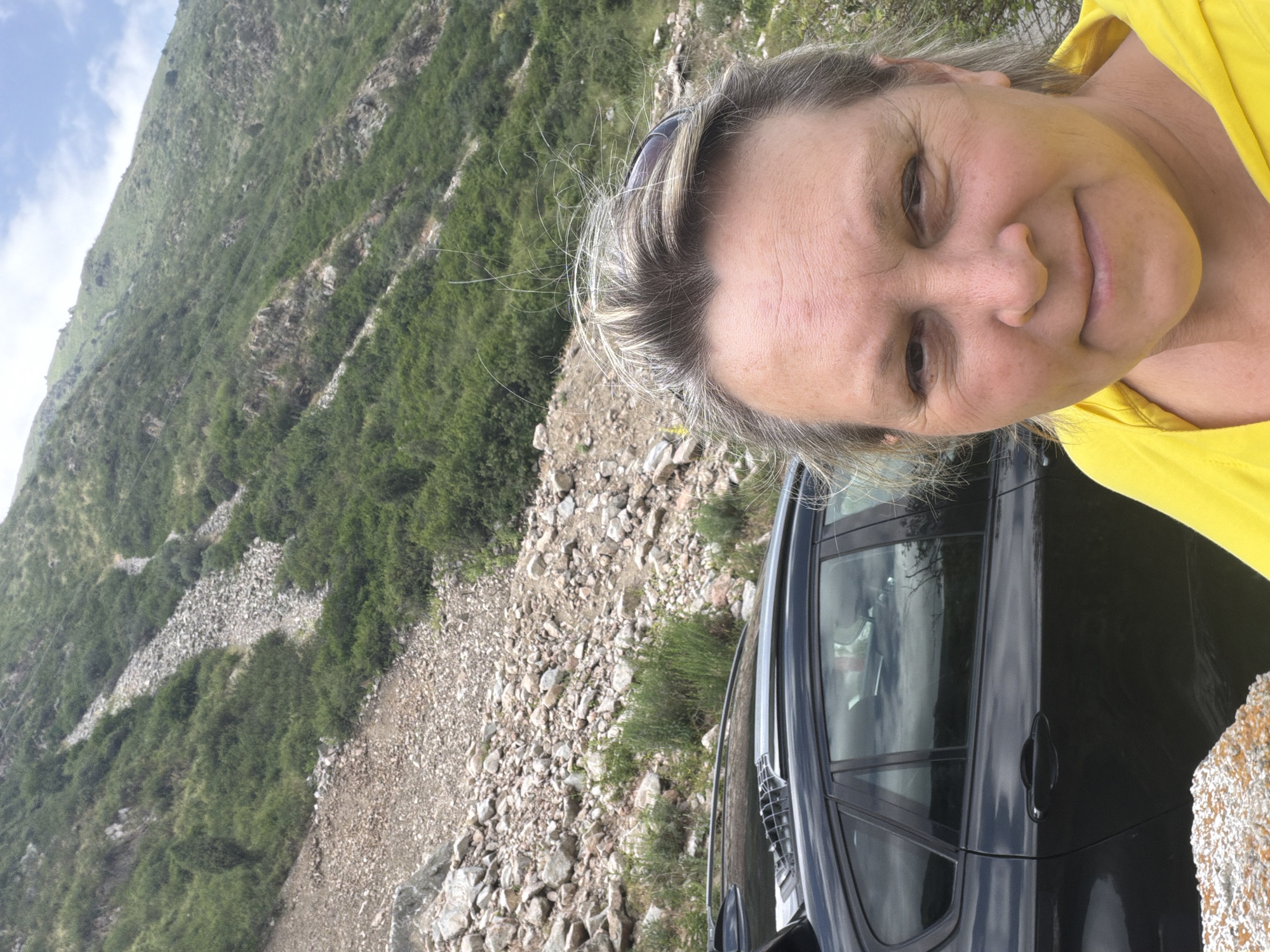 A woman with graying hair and a bright yellow hoodie smiles into the camera. She is standing in front of a black SUV, which is parked on a rocky, mountainous terrain. The background shows a steep, vegetated slope under a cloudy sky.