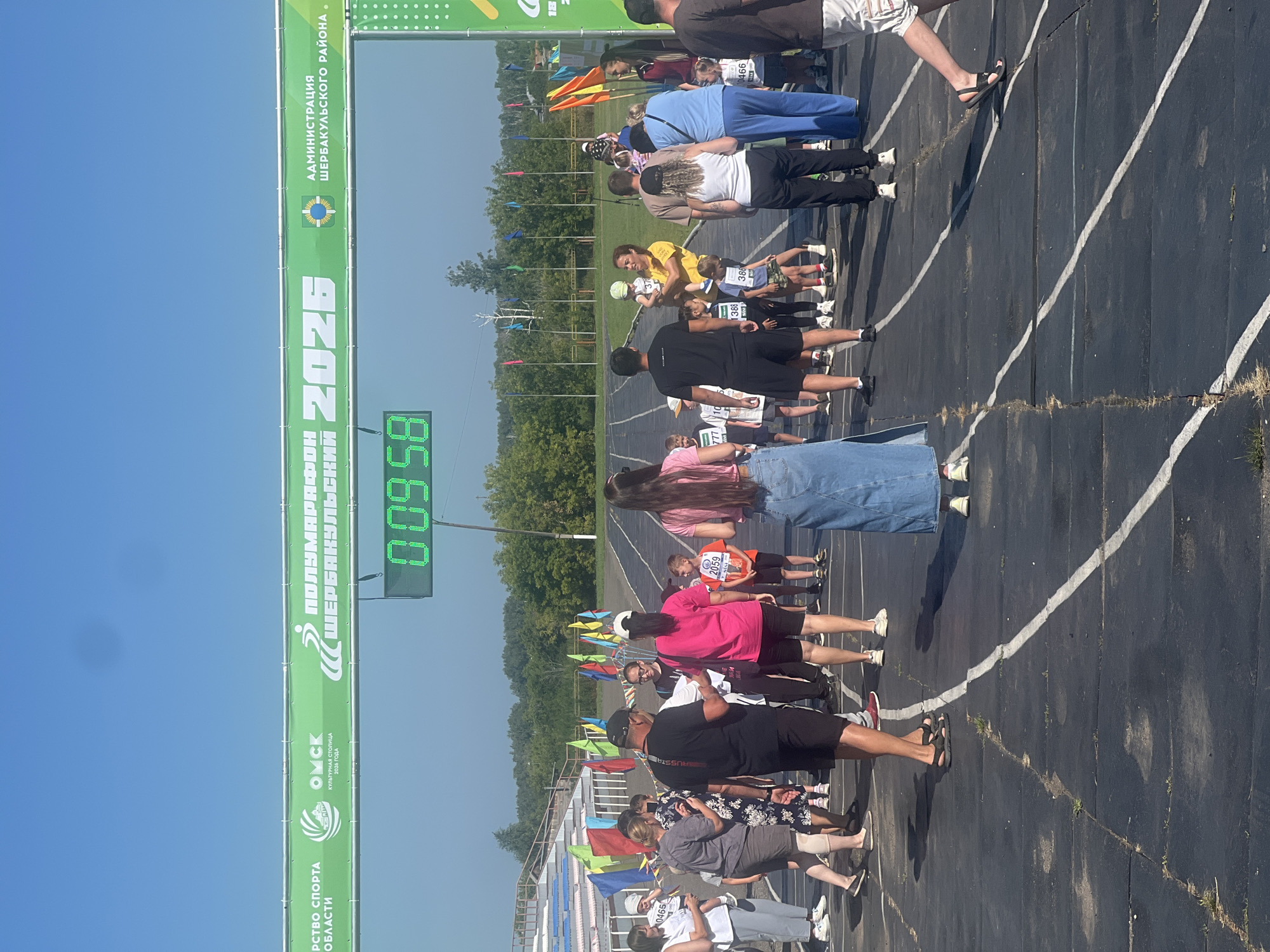 The image shows a group of people gathered on a dark asphalt track, likely a stadium or racecourse, under a clear blue sky. A green banner stretched across the top of the frame displays text in Russian, including "ПОЛУМАРАФОН" (half-marathon) and "ШЕРБАКУЛЬСКИЙ" (Sherbakulsky), indicating the location of the event is in Sherbakul, Russia. A digital timer displaying "00:09:58" is prominently visible, suggesting the start or a segment of a race.
Various individuals are present, some wearing athletic attire, others in casual clothing. Many appear to be participants, with some children in the foreground wearing bib numbers. There are also adults, some holding children or engaged in conversation. Flags of different colors are planted along the track, adding to the festive atmosphere of a sporting event. The background reveals a grandstand with blue and white seating, and a line of trees beyond the stadium. The ground surface is a dark asphalt track marked with white lines. The overall scene captures a moment at a community running event in Sherbakul, Russia.