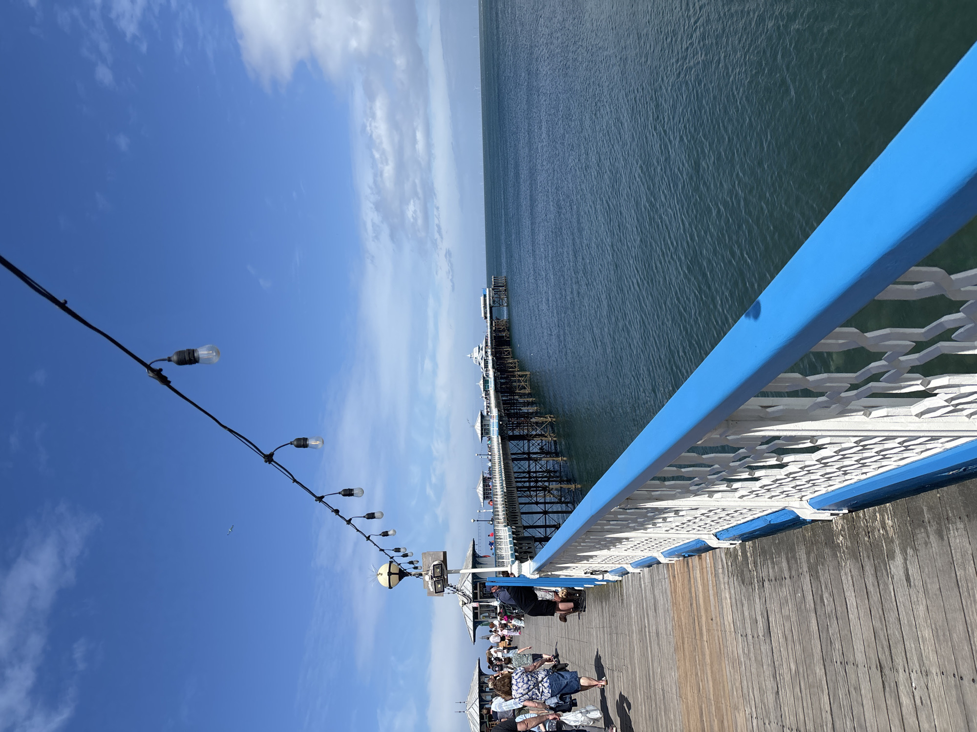 The image shows a view from Llandudno Pier in Llandudno, United Kingdom. The pier extends into the sea, with a blue and white railing visible along the right side of the frame. A string of lights hangs diagonally across the upper left of the image. People are visible walking and sitting along the pier. The sea is visible on the right, and the sky with scattered clouds is on the left.