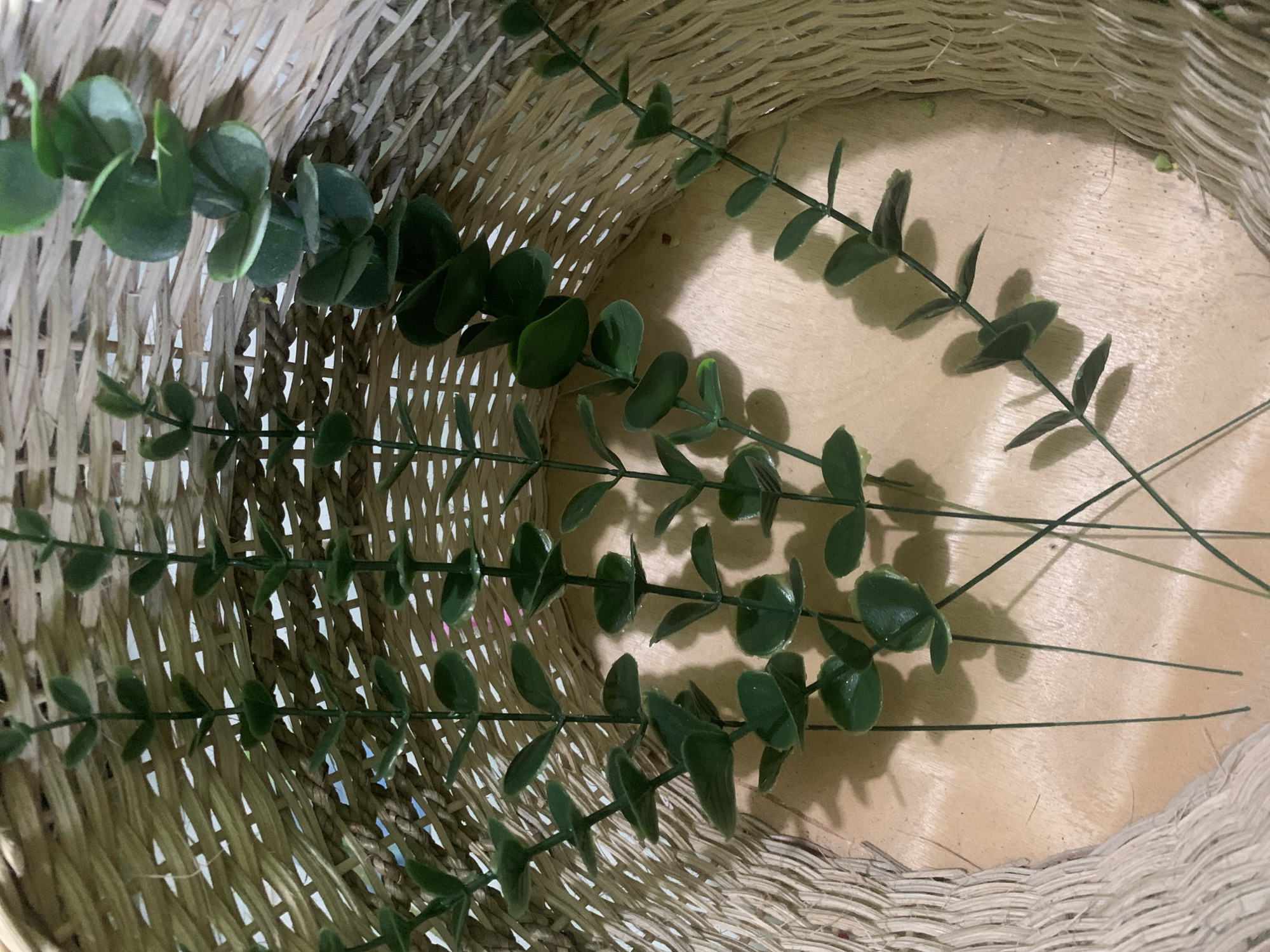 The image shows a close-up view of several artificial eucalyptus stems inside a woven basket. The basket is made of light-colored reeds and has a textured appearance. The eucalyptus stems are green with small, rounded leaves. Some of the stems are arranged in a way that suggests they are being placed or stored in the basket. The background is a light brown wooden surface. There are no people visible in the image, and no specific location in Kuala Pembuang, Indonesia, can be identified from the provided image. The scene is static and depicts a collection of decorative items.