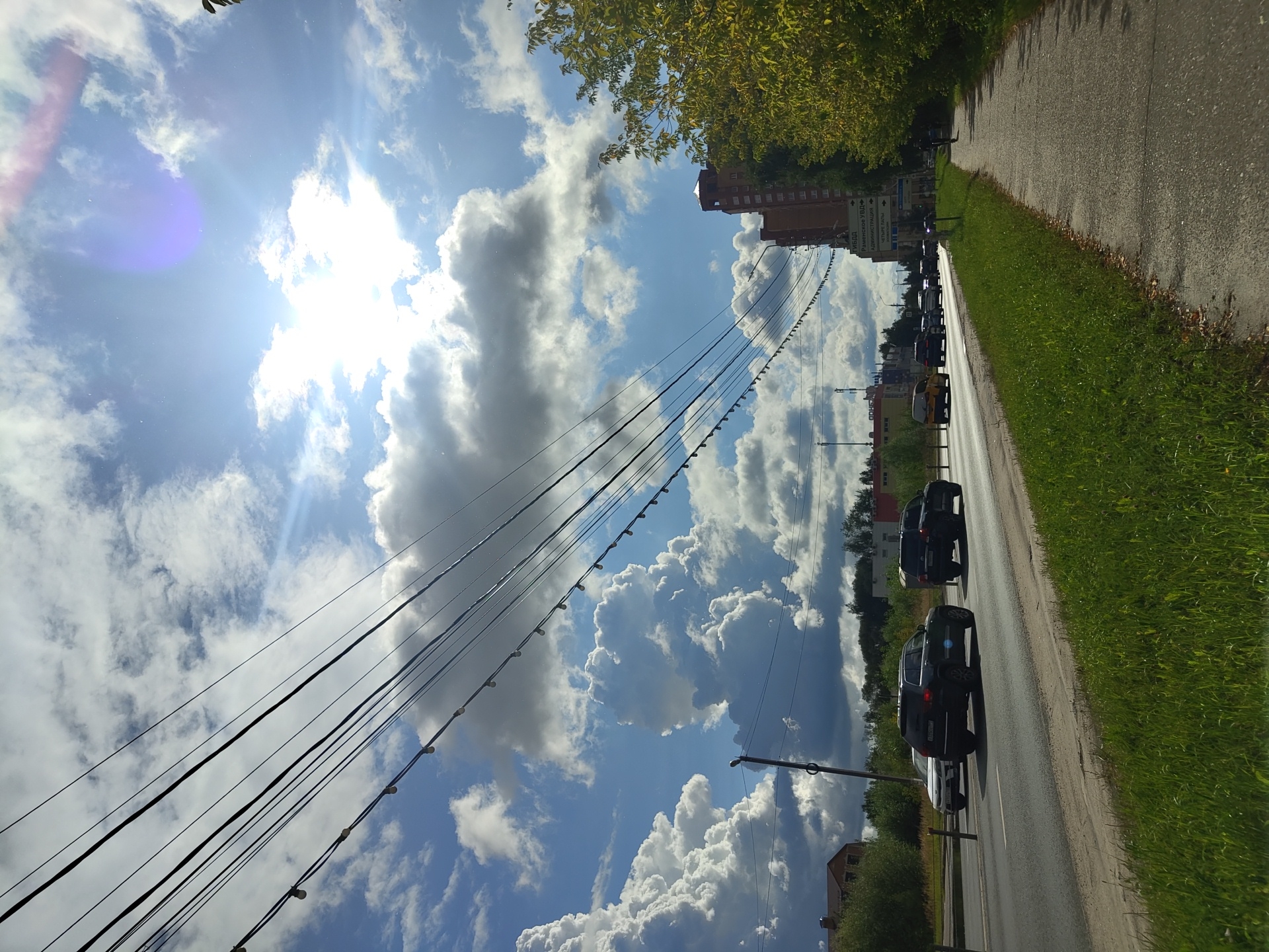 This is a vertical, eye-level, outdoor shot of a street scene in Ramenskoye, Russia, on a sunny day with scattered clouds. The perspective is from the side of the road, looking down the street. In the foreground, a paved sidewalk runs along the right side of the image, bordered by a strip of green grass. A large tree with yellowing leaves is on the far right, partially obscuring a tall brick building in the background. The street itself is paved and has multiple lanes of traffic moving from left to right. Several cars are visible on the road, including a dark SUV in the middle lane and a yellow car further ahead. Overhead, a tangle of power lines stretches across the frame, with small light bulbs strung between some of them, suggesting decorative lighting. The sky is a bright blue, filled with large, puffy white and gray clouds, with the sun prominently visible, casting a lens flare and a purplish haze. In the distance, more buildings and trees can be seen, along with a street sign that appears to be in Russian. The overall atmosphere is that of a typical urban street on a bright, partly cloudy day.