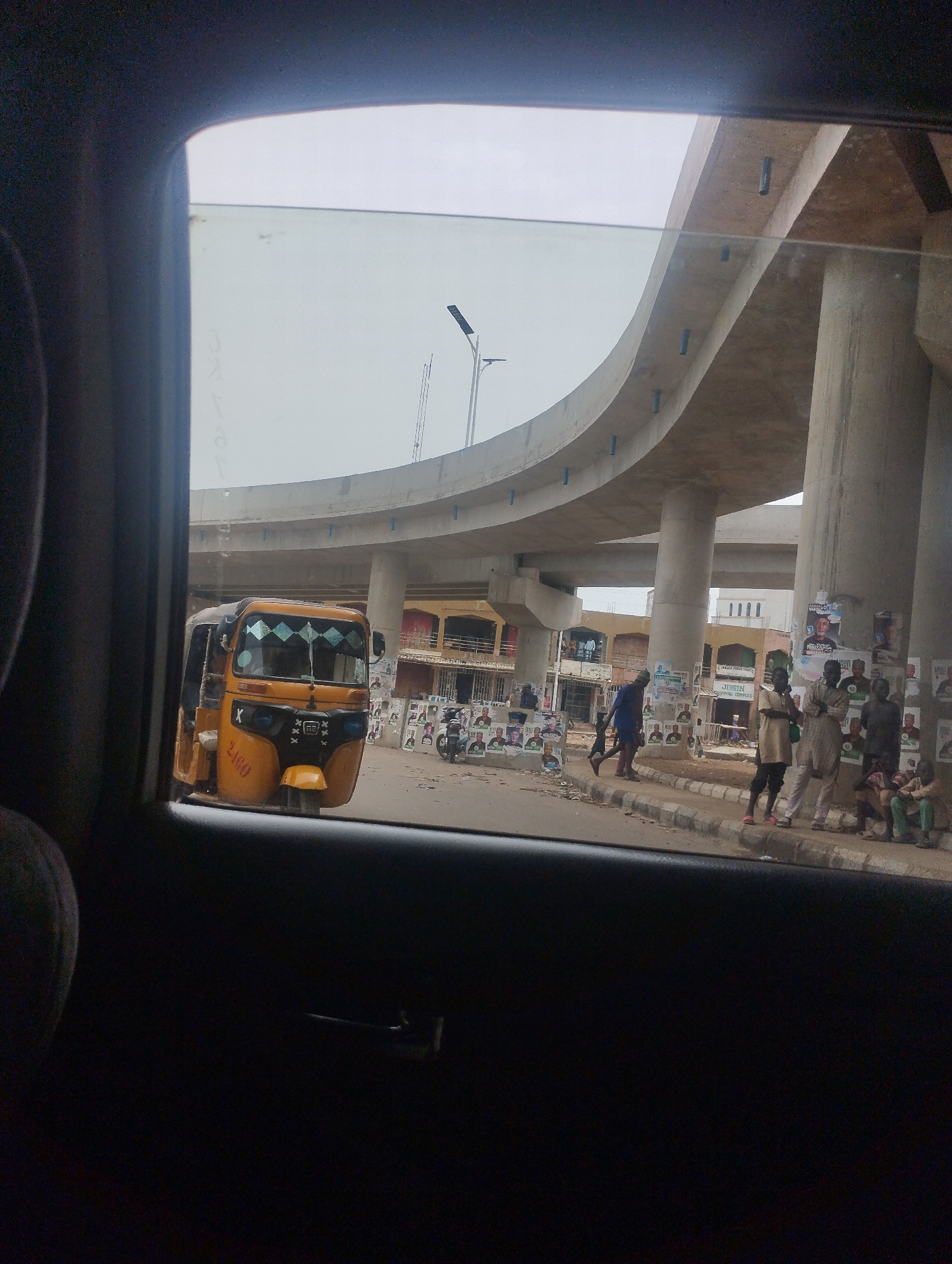 The image, taken from inside a vehicle through a window, shows a street scene in Bauchi, Nigeria. A yellow tricycle ("Keke Napep") is parked on the side of the road. An elevated concrete highway overpass dominates the upper portion of the view. Along the sidewalk, several people are standing and sitting, near buildings and posters affixed to the columns of the overpass. A man is walking across the sidewalk, carrying something in his left hand. The weather appears overcast.